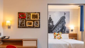 Una camera d'albergo con una scrivania, una sedia rossa, un letto con biancheria bianca, un cuscino colorato e un muro con quadri.