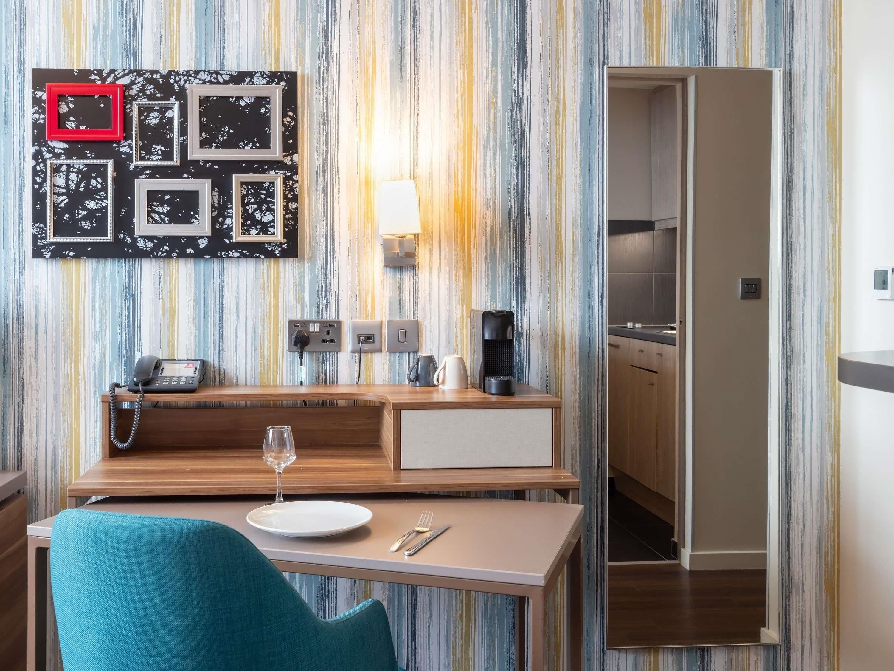 A modern hotel room with a striped wall, a wooden desk with a phone and a coffee maker, a glass of wine, and a teal chair.
