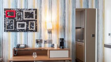 A modern hotel room with a striped wall, a wooden desk with a phone and a coffee maker, a glass of wine, and a teal chair.