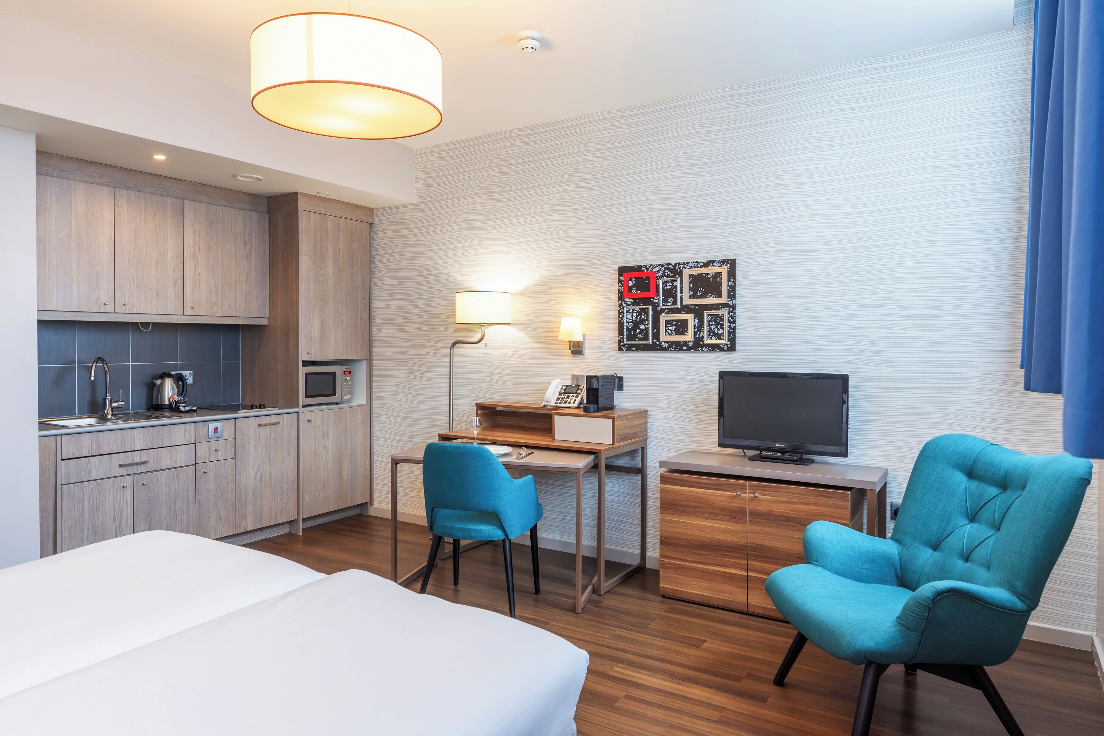 A hotel room with a bed, a desk with a chair, a television, a kitchenette, and a wall-mounted artwork.