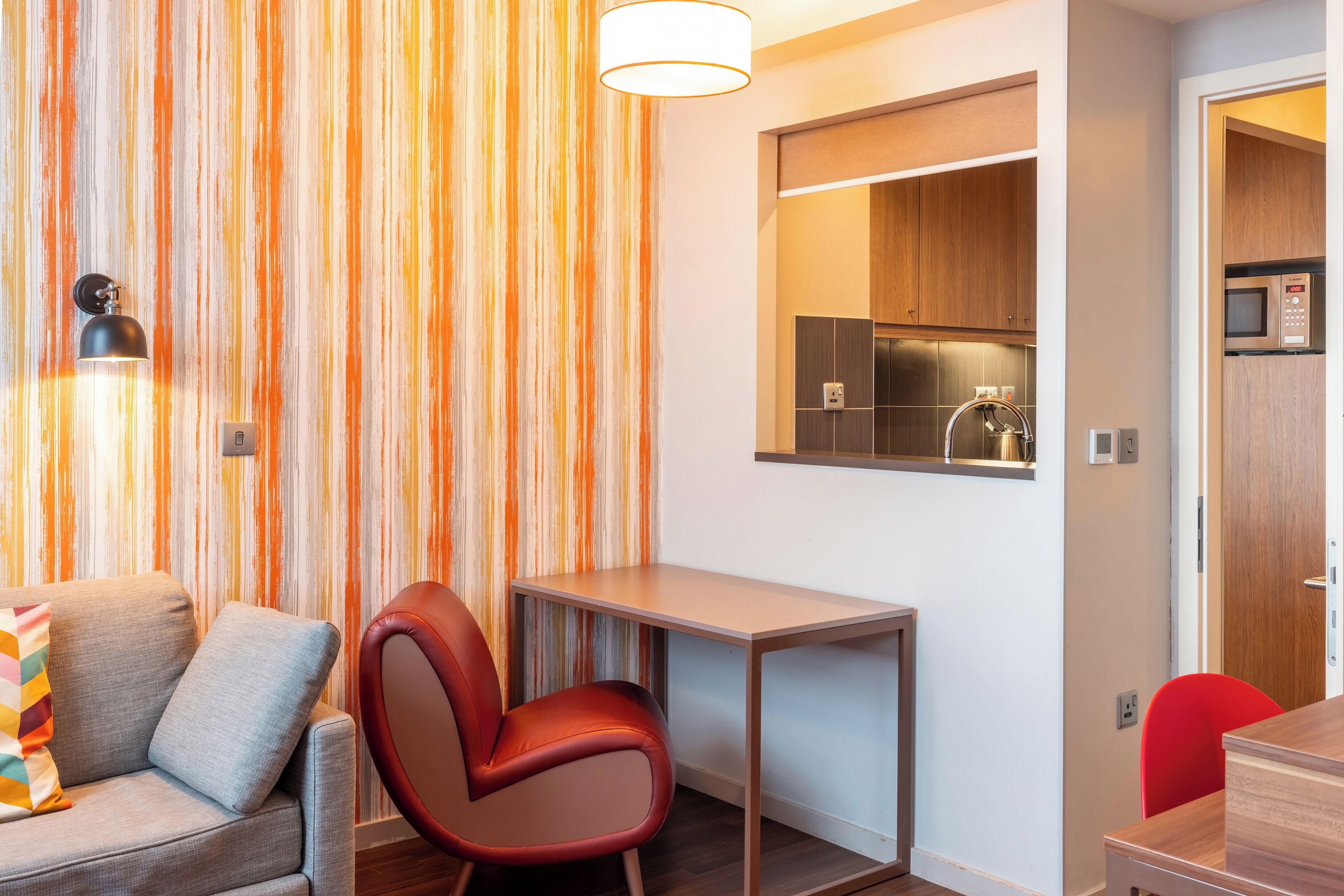 A compact living space with a sofa, a red chair, a small table, and a kitchen area with a microwave and a kettle.