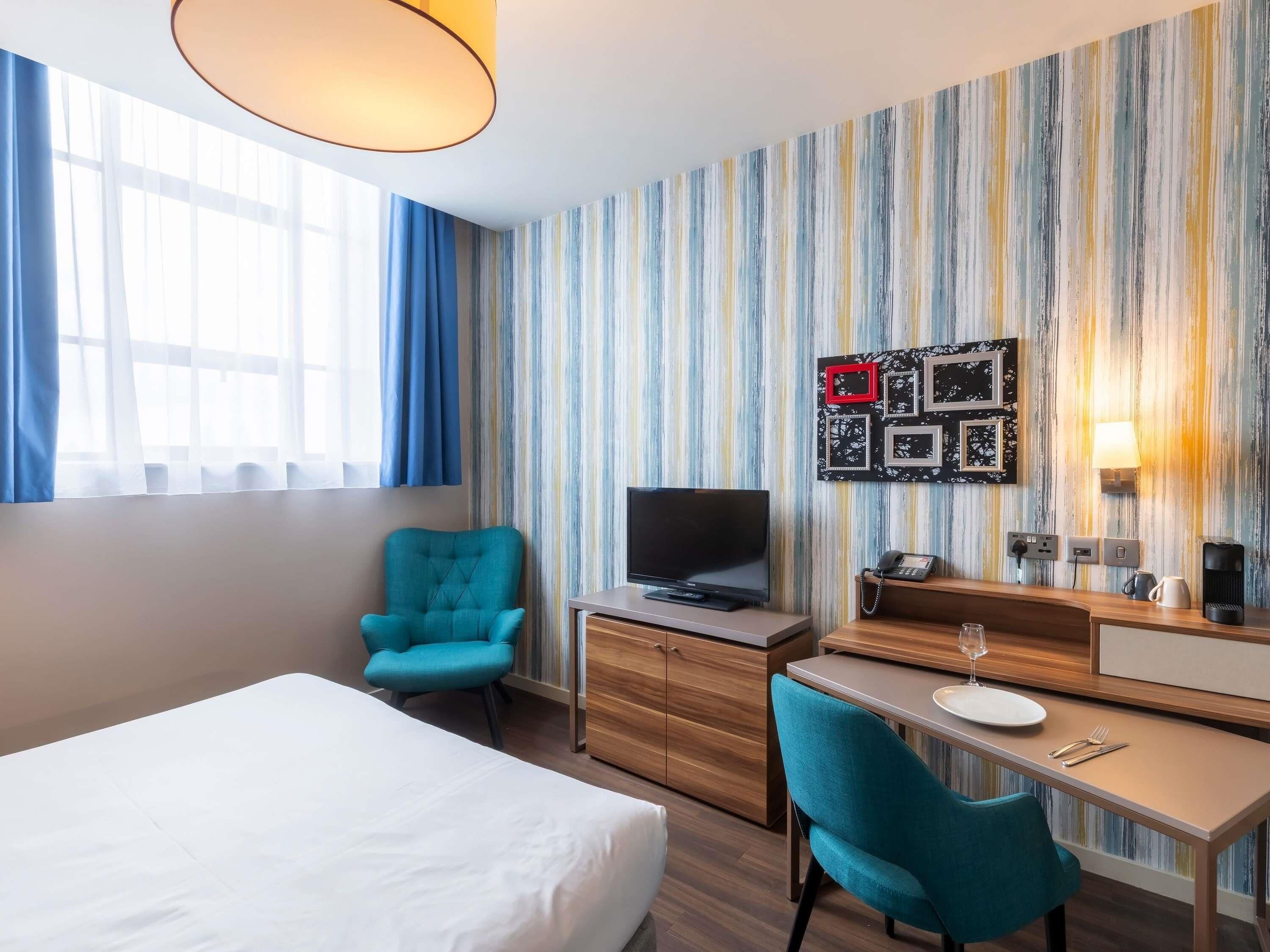 A hotel room with a bed, a desk, a television, a chair, a table with a plate and cutlery, a coffee maker, and a wall with a striped pattern.
