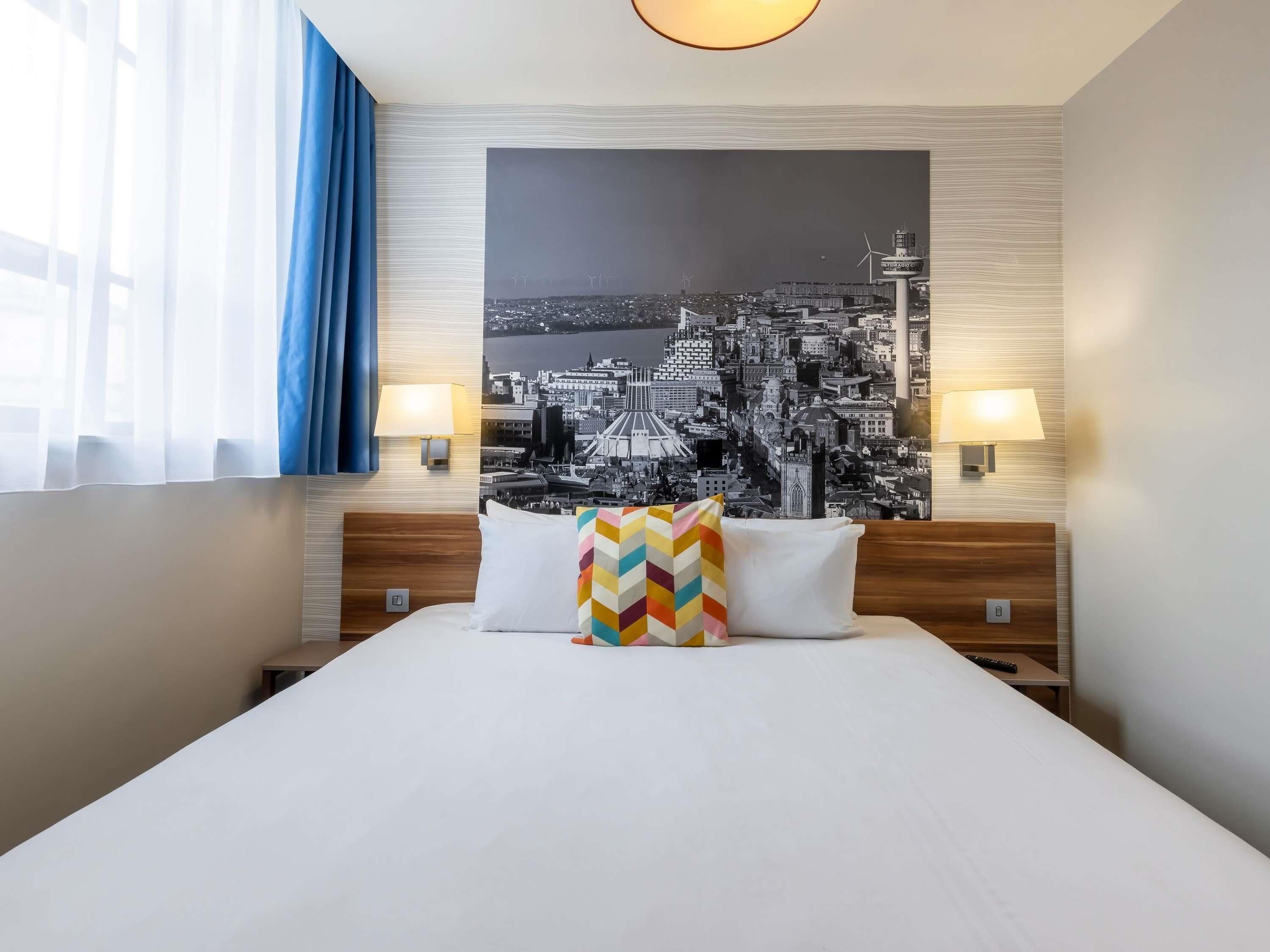 A hotel room with a bed, two bedside lamps, a cityscape wall art, and a window with curtains.