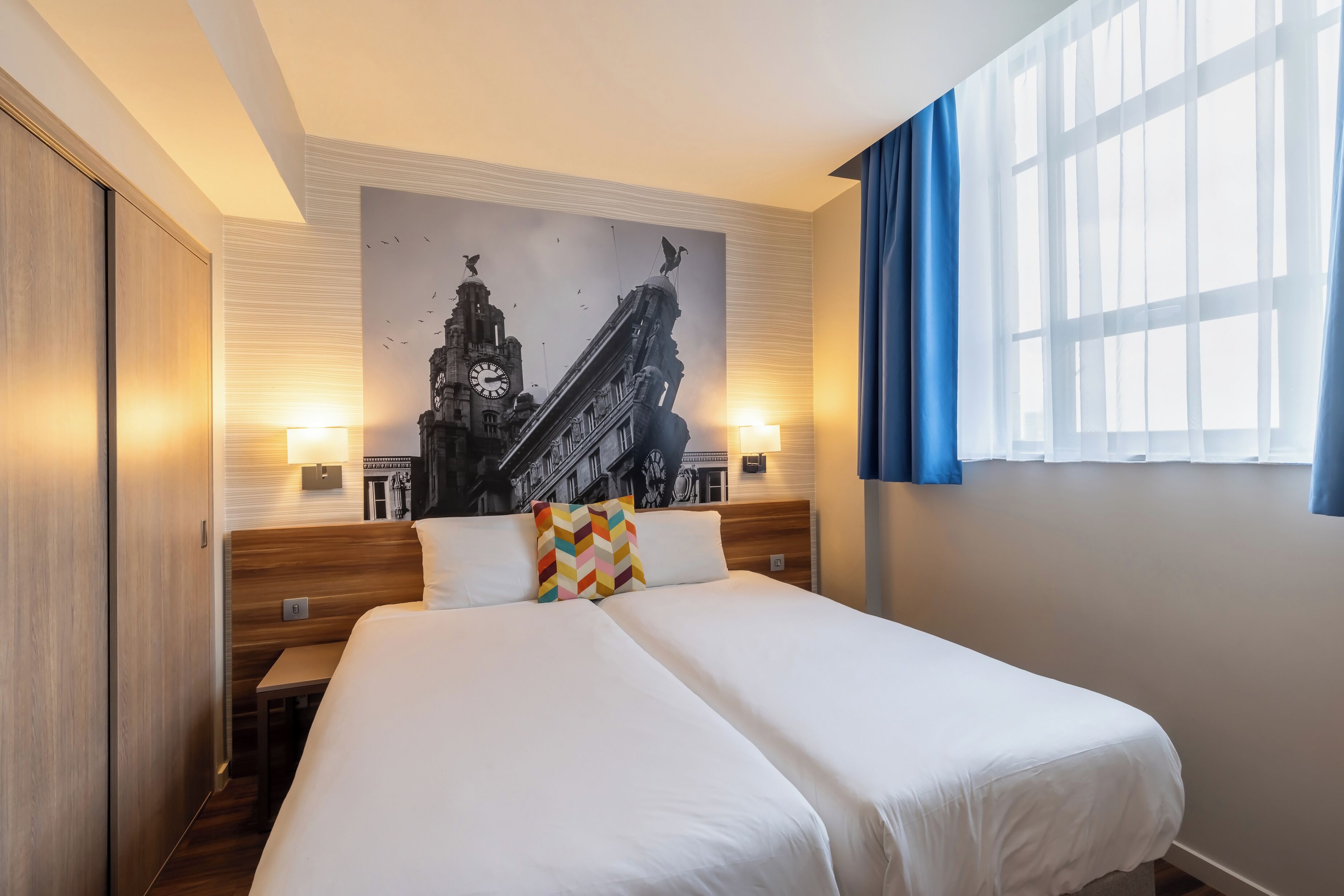 A hotel room with a large bed, a headboard with a cityscape mural, two bedside tables with lamps, and a window with blue curtains.