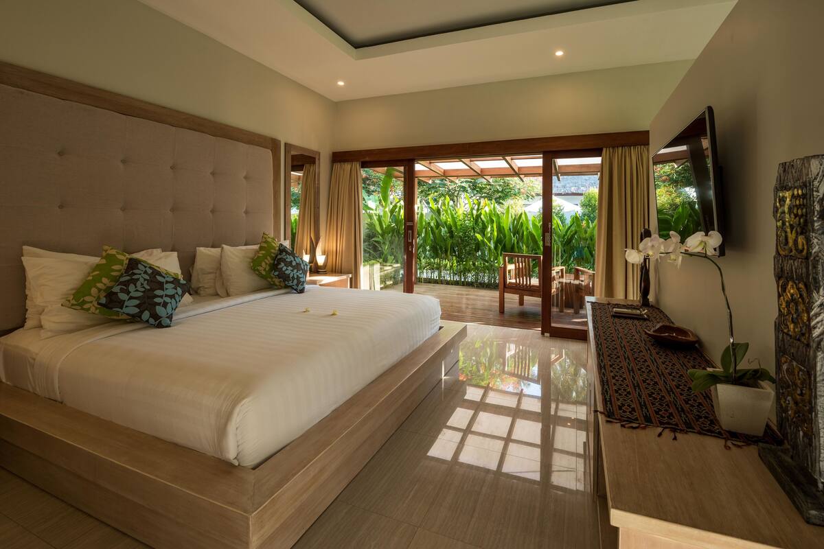 A modern bedroom with a large bed, a TV, and a view of greenery outside.