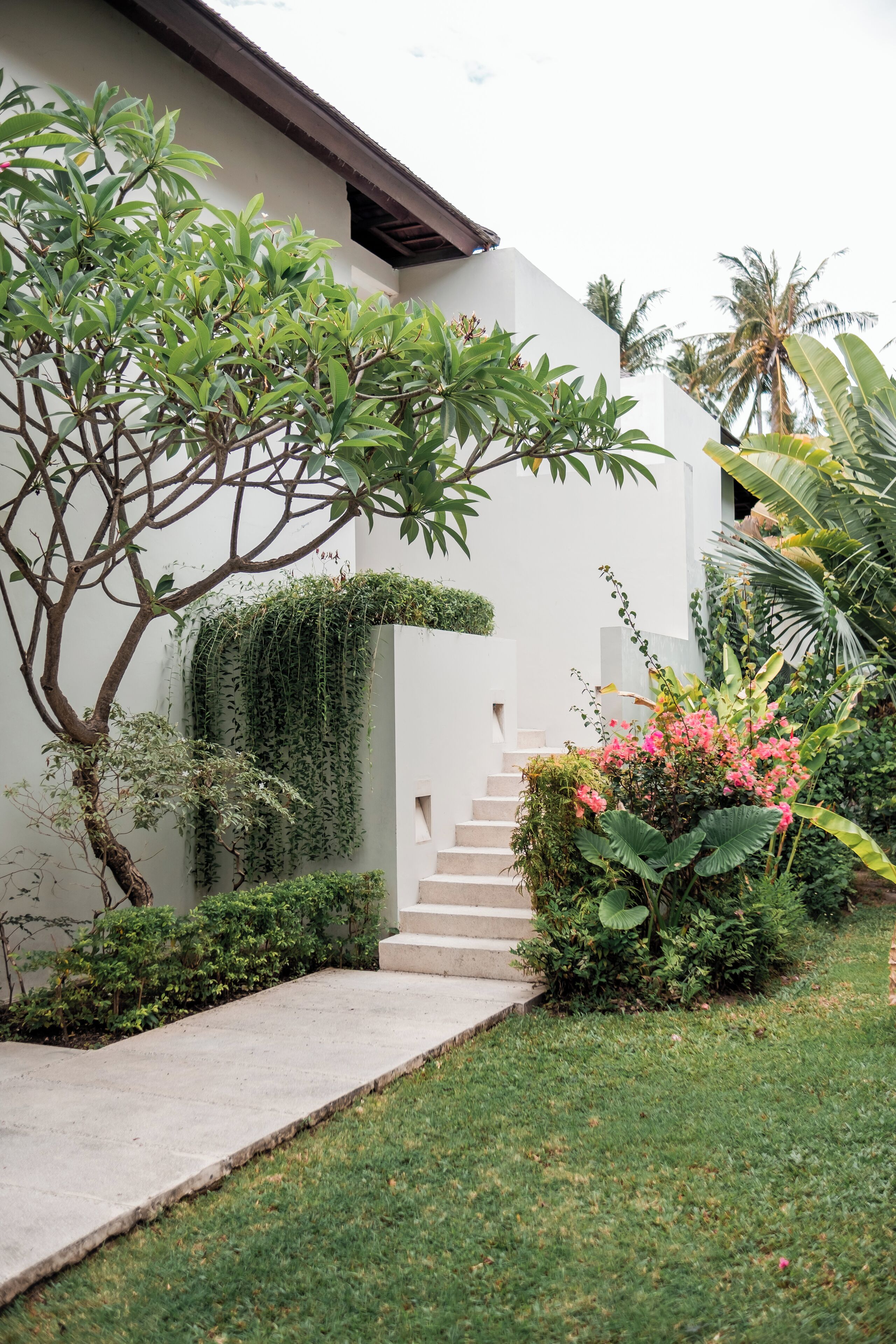A white building with a staircase leading up to a door, surrounded by greenery and a small garden.