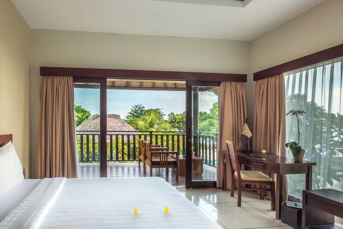 A hotel room with a bed, a desk with a TV, a chair, and a balcony with a view of greenery and a thatched-roof structure.