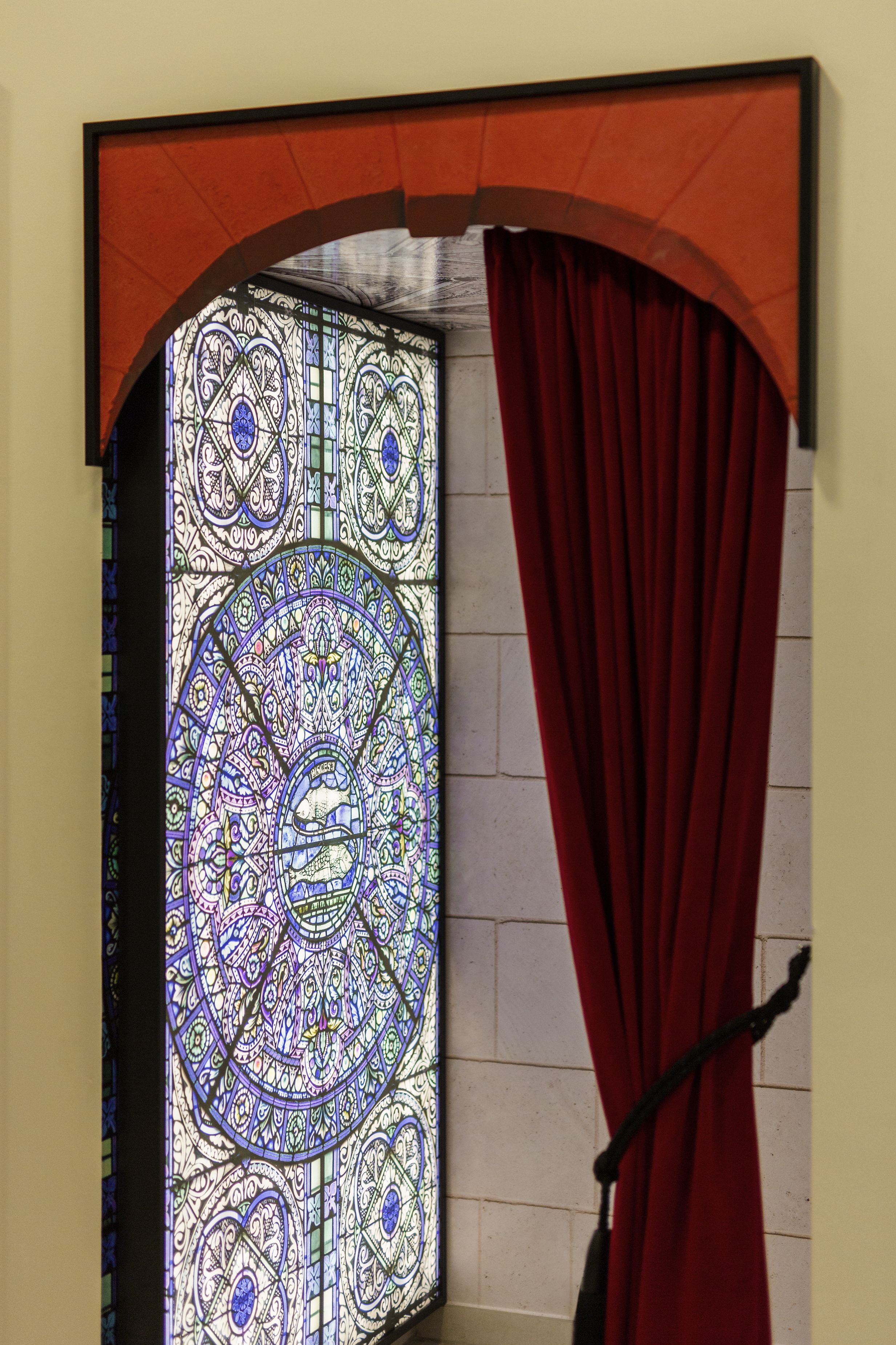 Une fenêtre en vitrail aux motifs complexes et un rideau rouge, partiellement tiré sur le côté.