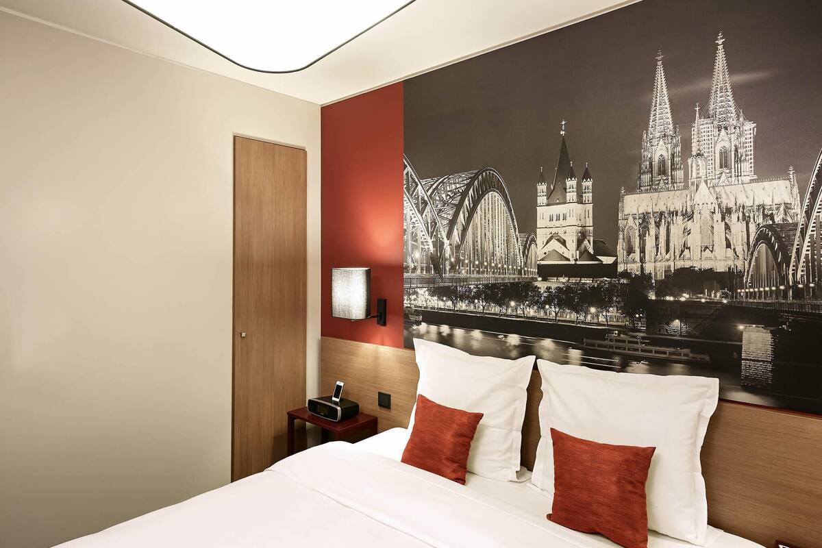 Quarto de hotel com cama, mesa de cabeceira, luminária e um grande mural da paisagem urbana na parede.