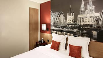 Una habitació d'hotel amb un llit, una taula de nit, una làmpada i un mural gran amb un paisatge urbà a la paret.