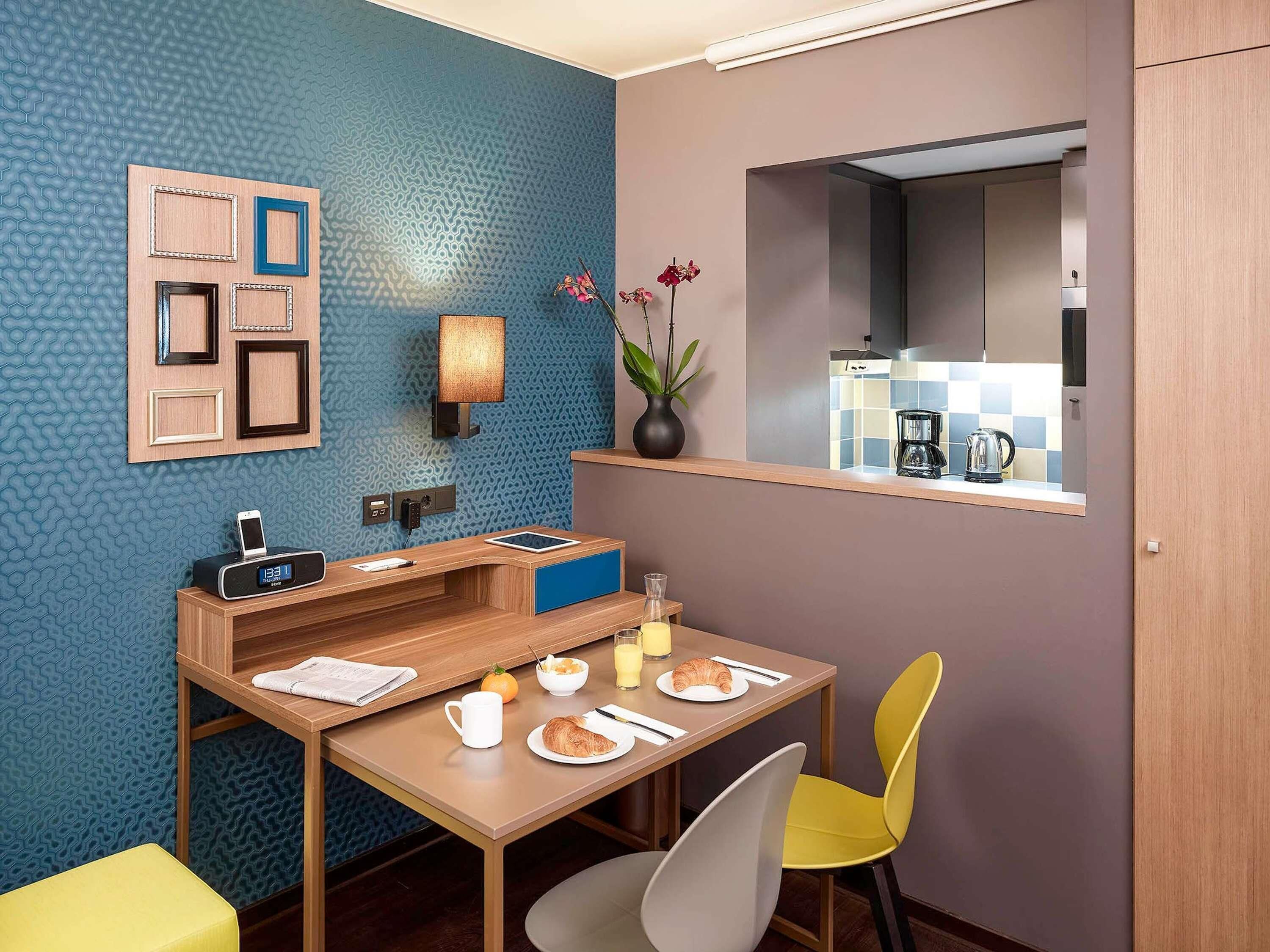 Une chambre d’hôtel compacte comprenant une table pour le petit-déjeuner, un petit coin salon, une horloge murale, un vase avec des fleurs et une vue sur la cuisine par une fenêtre.