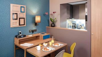 A compact hotel room with a breakfast table, a small seating area, a wall-mounted clock, a vase with flowers, and a view of the kitchen through a window.