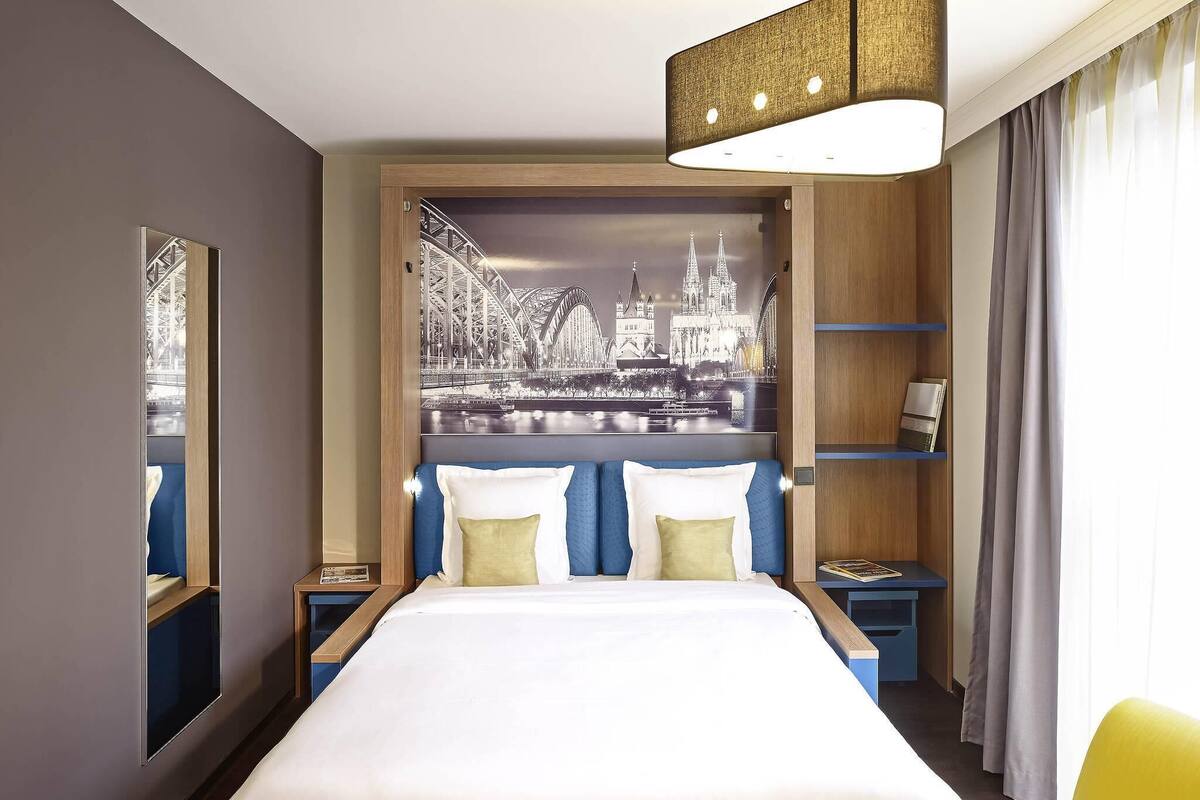 Quarto de hotel com uma cama grande, um mural com vista para a cidade, uma estante e uma mesa com abajur.