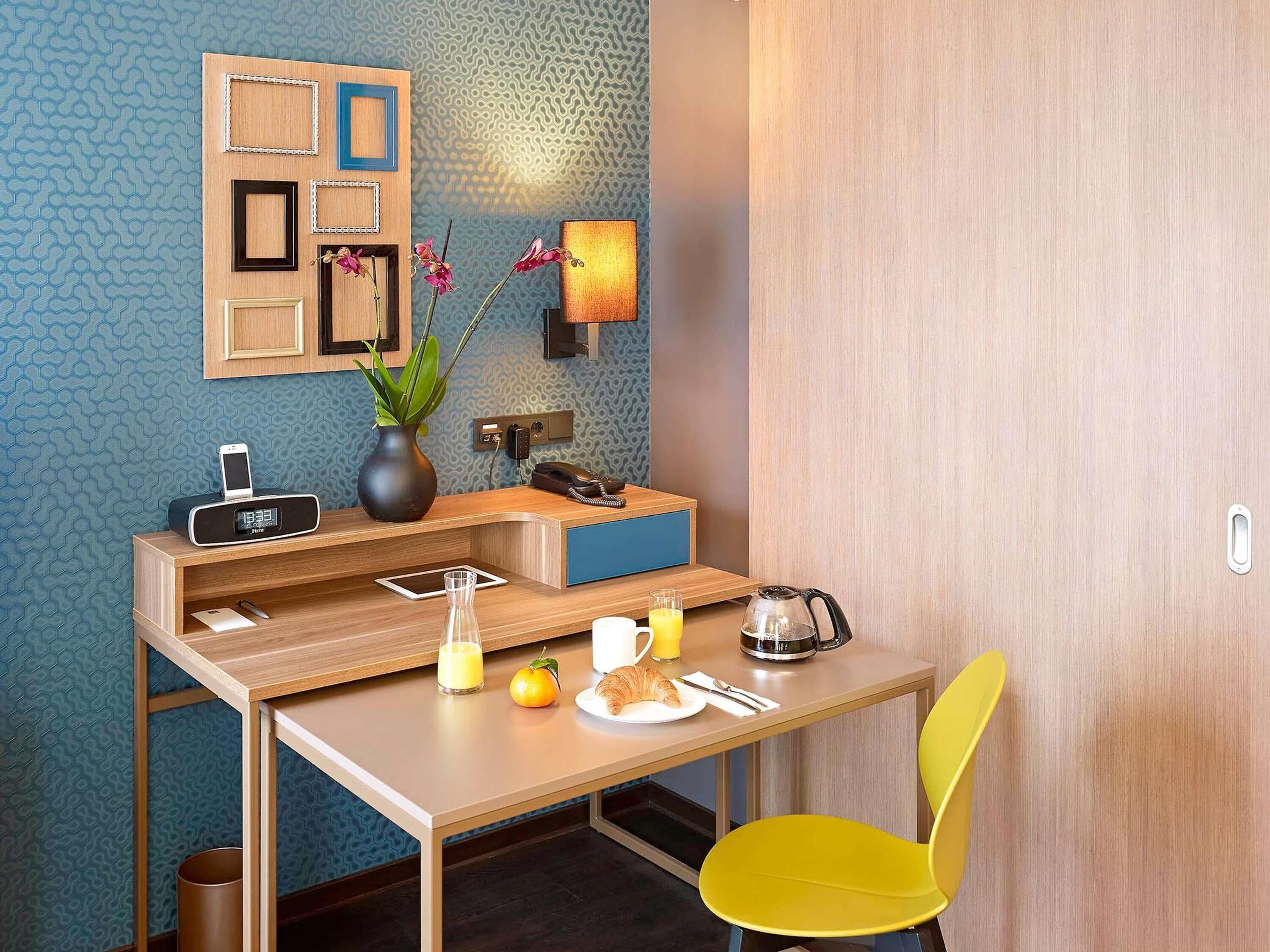 A compact hotel room with a desk, chair, and a small table with breakfast items.