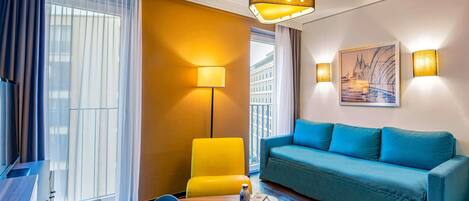 Une chambre d’hôtel avec un canapé bleu, une chaise jaune, une table ronde sur laquelle se trouvent un livre et une bouteille, un lampadaire et un tableau accroché au mur.