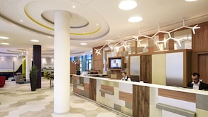 A modern reception area with a wooden reception desk, a large potted plant, and a digital screen displaying information. - Aparthotel Adagio Köln City (Cologne)