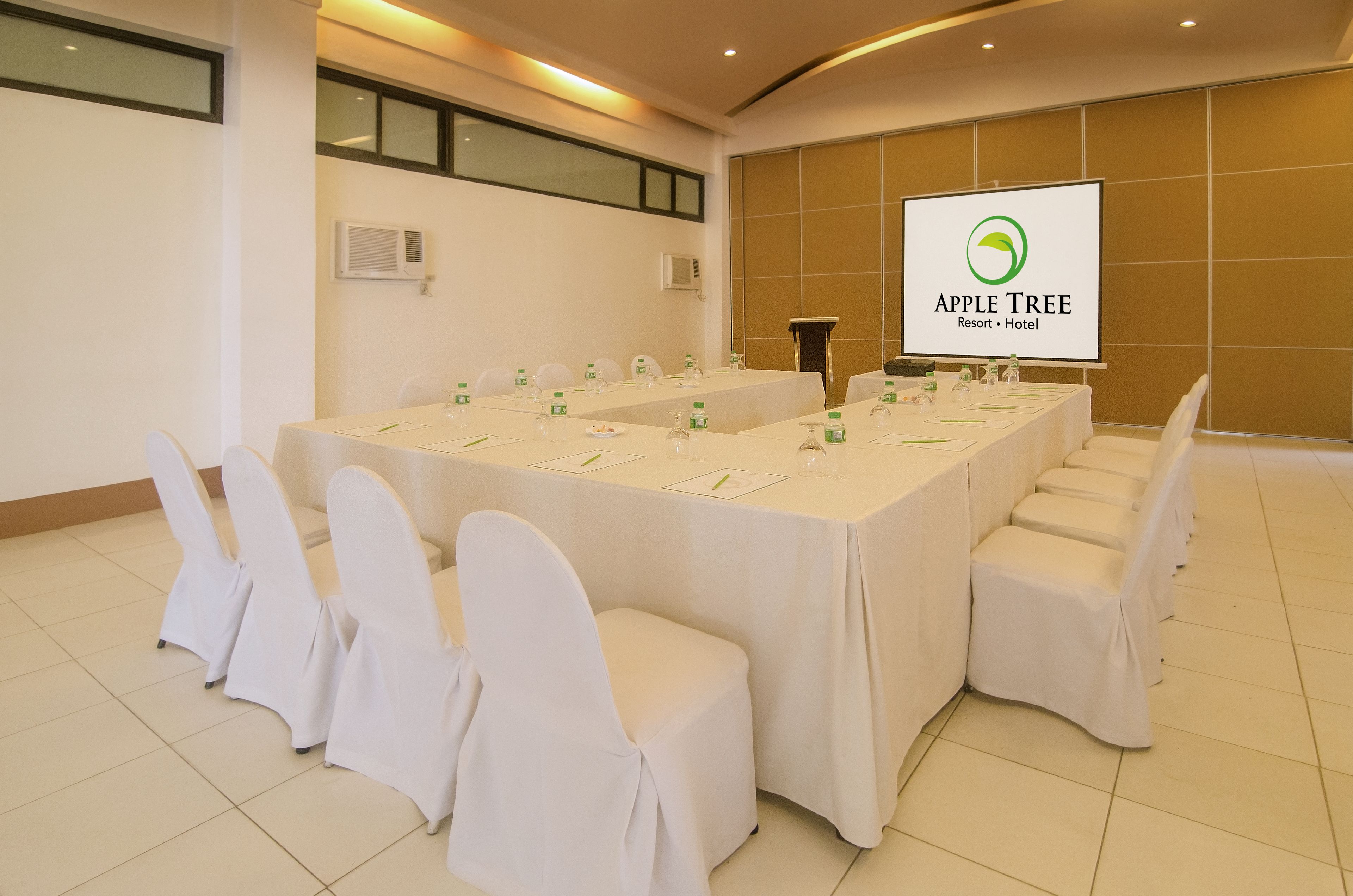 A conference room with a long table, white chairs, and a screen displaying the Apple Tree Resort Hotel logo.