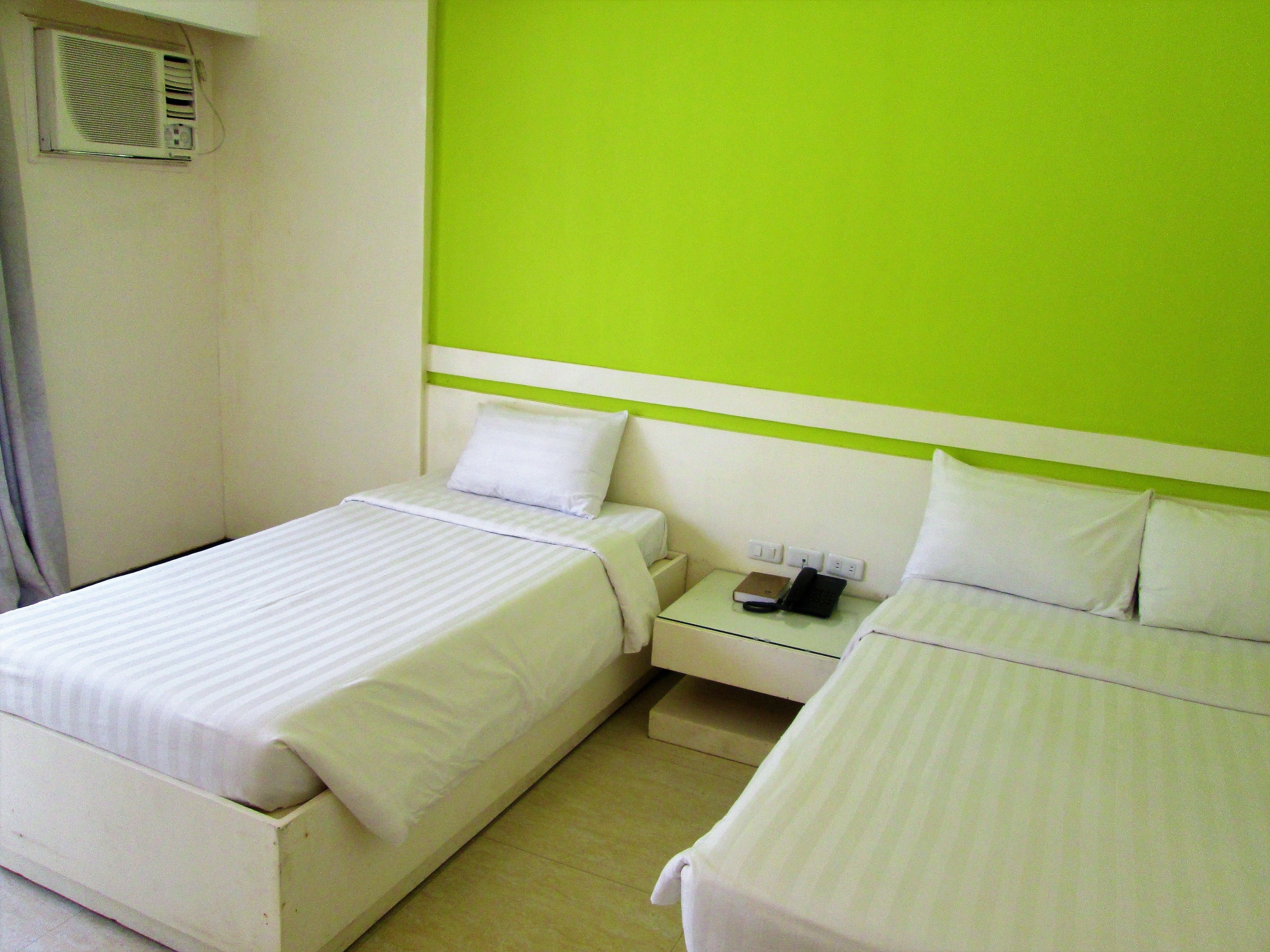 Habitación de hotel con dos camas, una mesita de noche con un teléfono y un libro, y un aire acondicionado instalado en la pared.