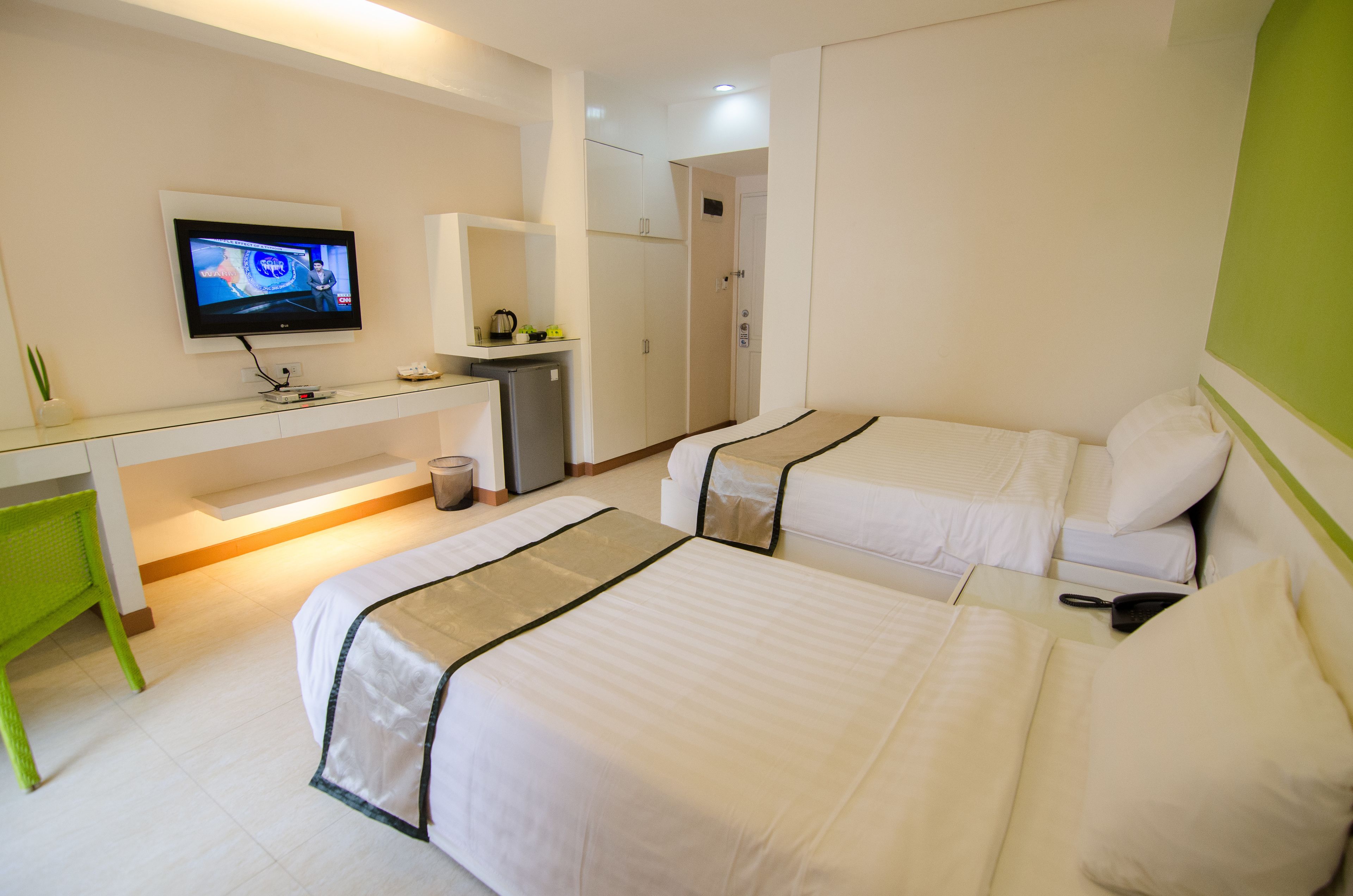 A hotel room with two beds, a desk, a television, and a small kitchenette.