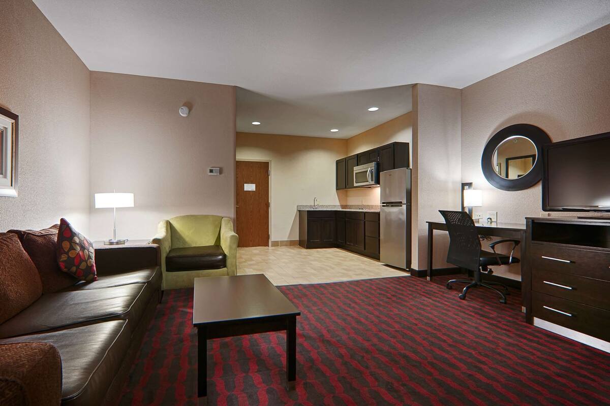 A hotel room with a sofa, armchair, dining table, kitchenette, and a TV.