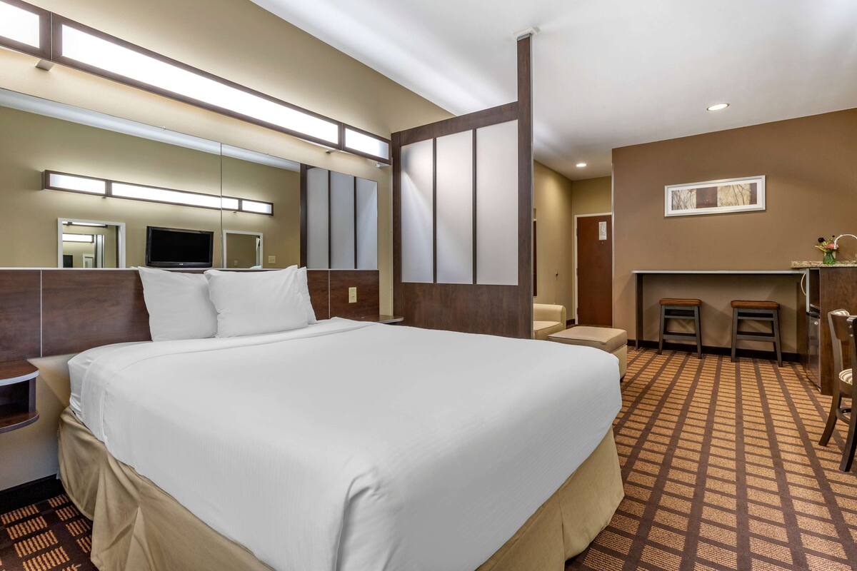A hotel room with a large bed, a TV, a mirror, a small seating area, and a kitchenette.