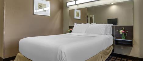 A hotel room with a large bed, a bedside table with a vase of flowers, a flat-screen TV, and a large mirror above the bed.