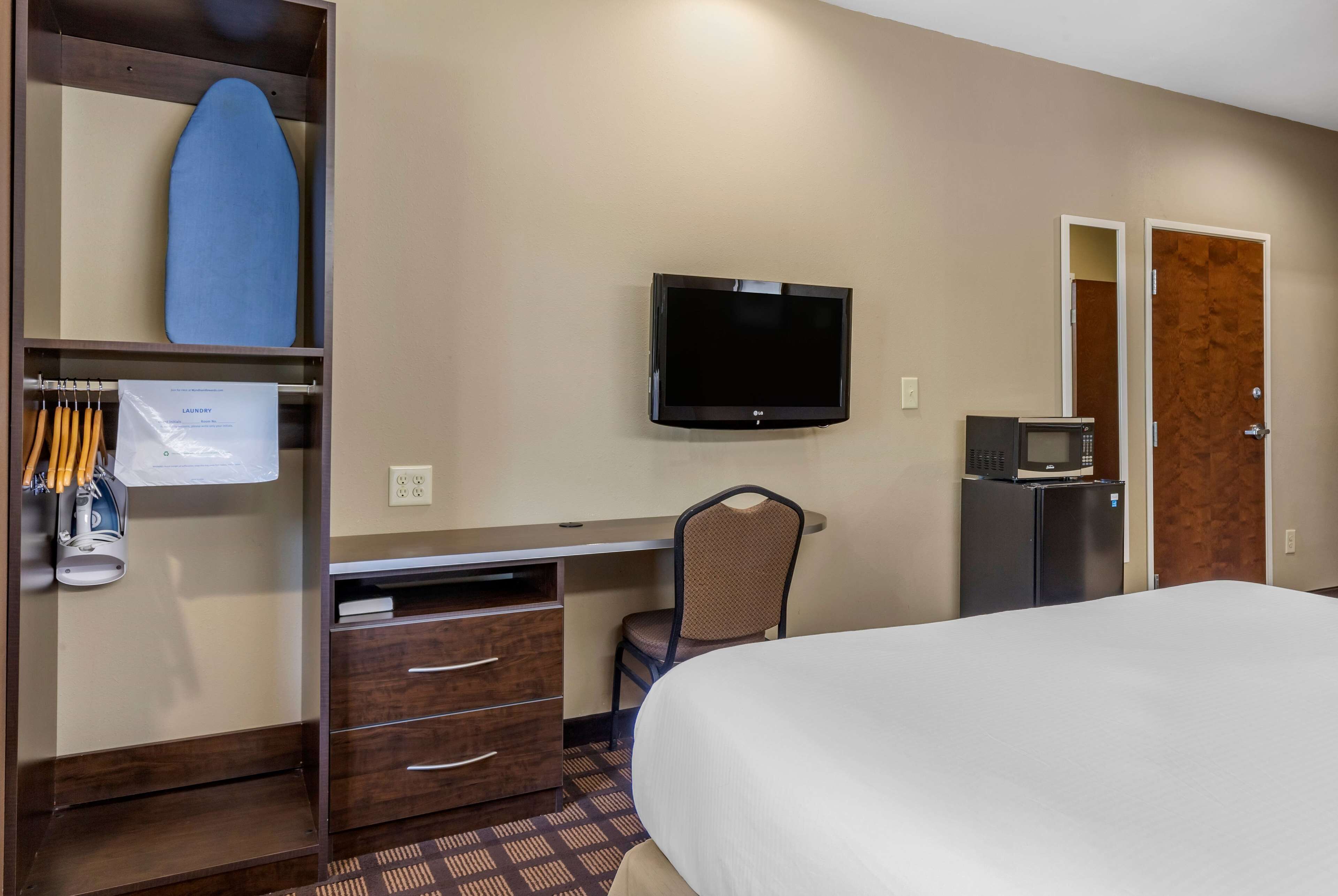 Une chambre d’hôtel comprenant un lit, un bureau avec des tiroirs, une chaise, une télévision, un four à micro-ondes et une armoire contenant une planche de surf et un sac à linge.