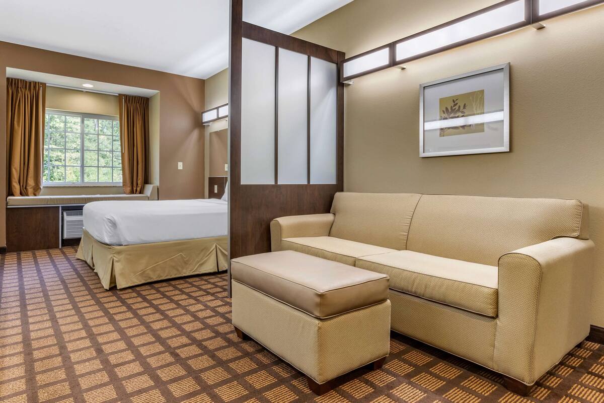 A hotel room with a bed, a sofa, an ottoman, a window with curtains, a wall-mounted artwork, and a patterned carpet.