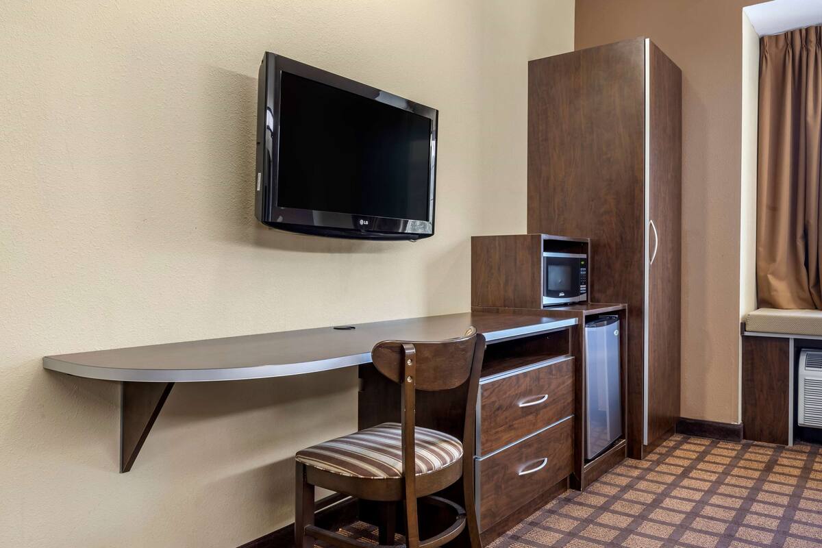 A compact hotel room with a flat-screen TV mounted on the wall, a small kitchenette with a microwave, a refrigerator, and a wooden desk with a chair.