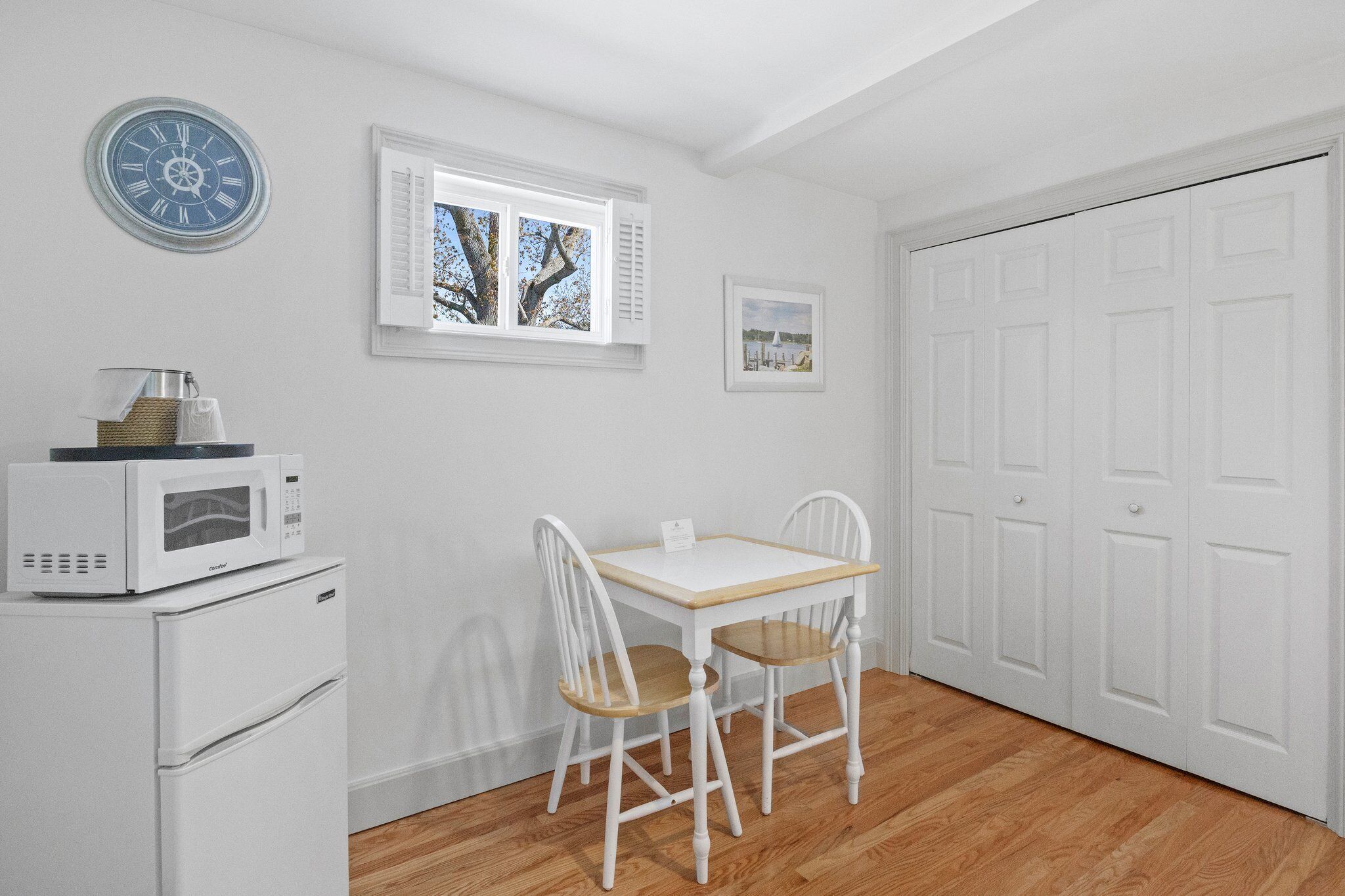 A small, white room with a wooden floor, a white table with two chairs, a white refrigerator, a microwave, a window with shutters, and a white door.