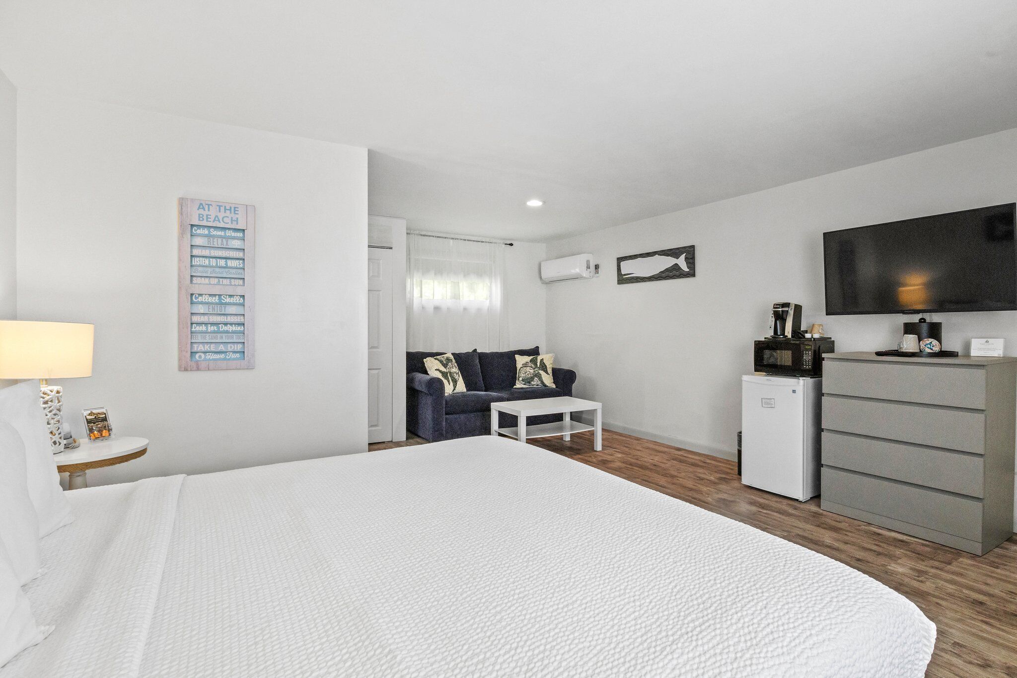 A modern hotel room with a large bed, a small desk, a sofa, a TV, and a mini-fridge.