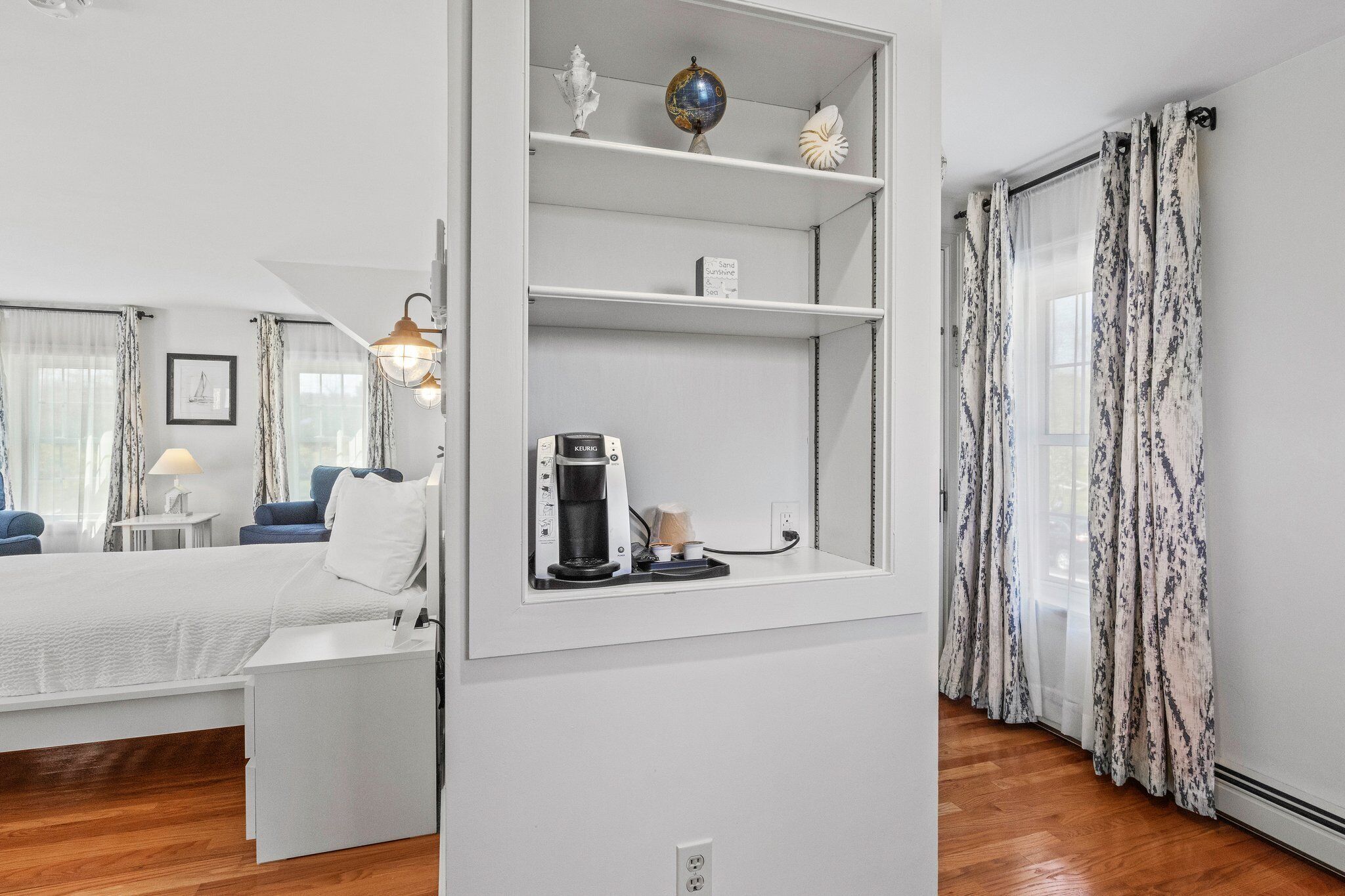 A modern hotel room with a bed, a coffee machine, a shelf with decorative items, and a view of the outdoors through sheer curtains.