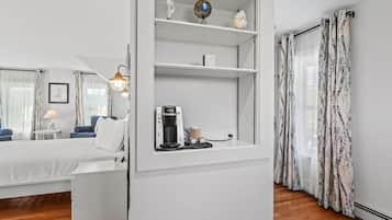 A modern hotel room with a bed, a coffee machine, a shelf with decorative items, and a view of the outdoors through sheer curtains.