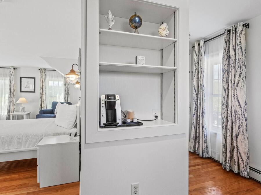 A modern hotel room with a bed, a coffee machine, a shelf with decorative items, and a view of the outdoors through sheer curtains.