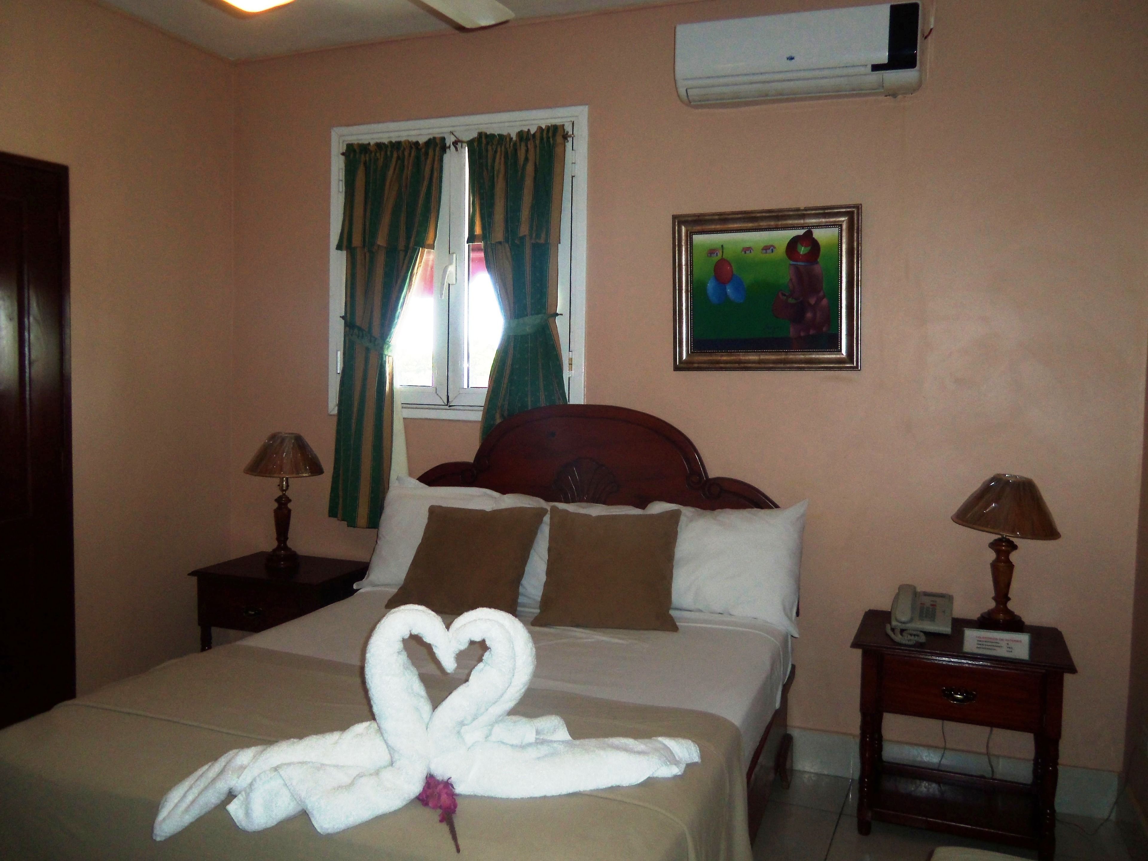 A hotel room with a bed, a nightstand, a bedside table, a window with curtains, an air conditioner, and a painting on the wall.