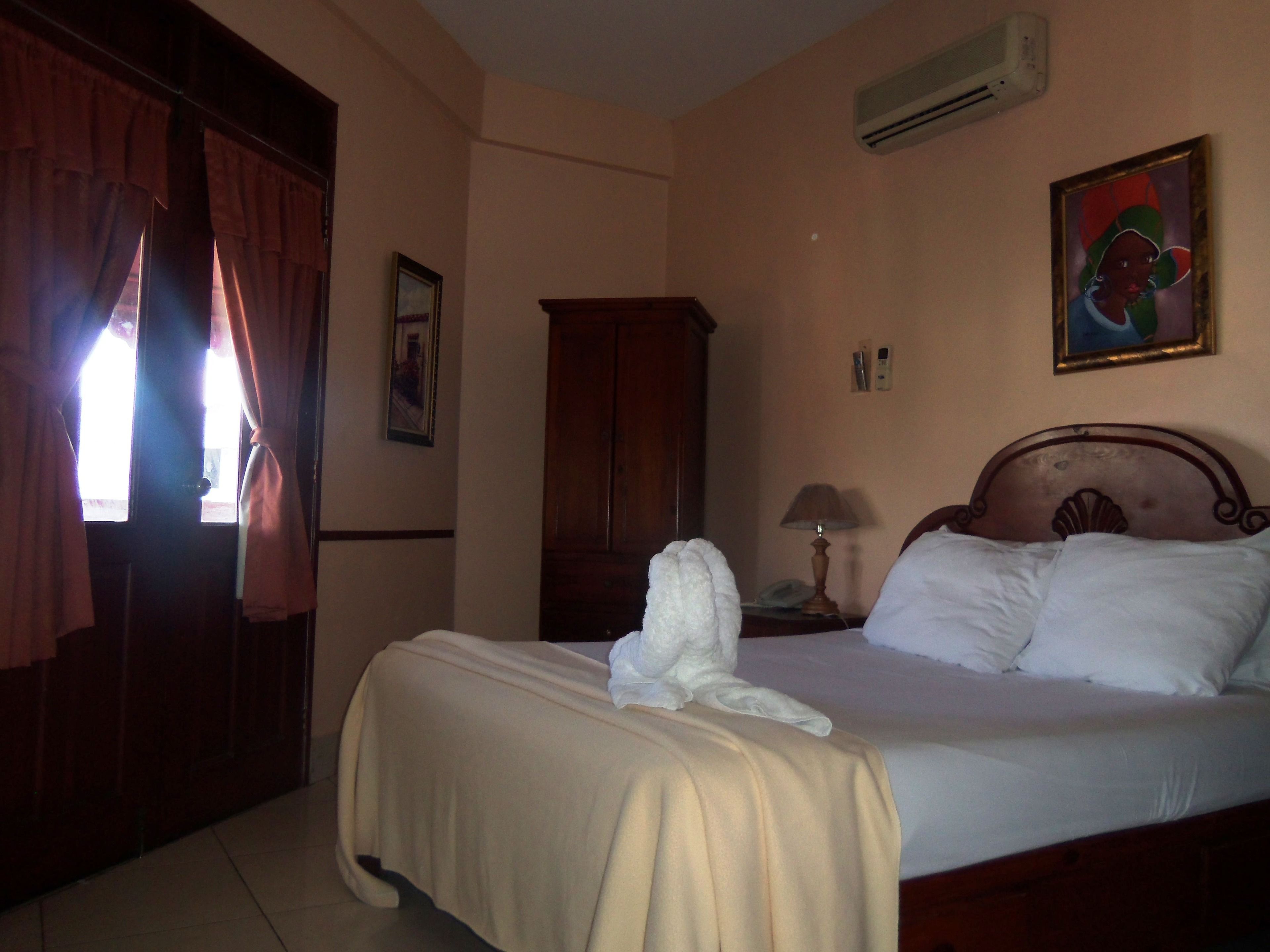 A hotel room with a bed, a wooden wardrobe, a painting on the wall, and an air conditioning unit.