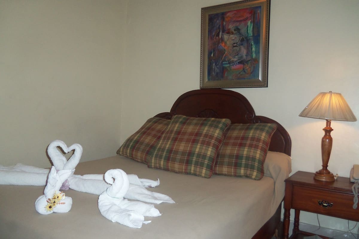 A neatly made bed with a heart-shaped towel arrangement, plaid pillows, and a bedside lamp.