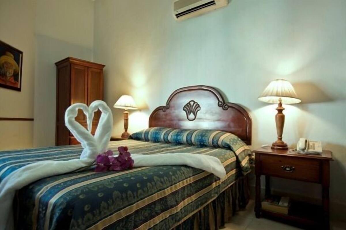 A neatly made bed with a heart-shaped towel and purple flowers, two bedside lamps, a wooden nightstand, and a wall-mounted air conditioner.