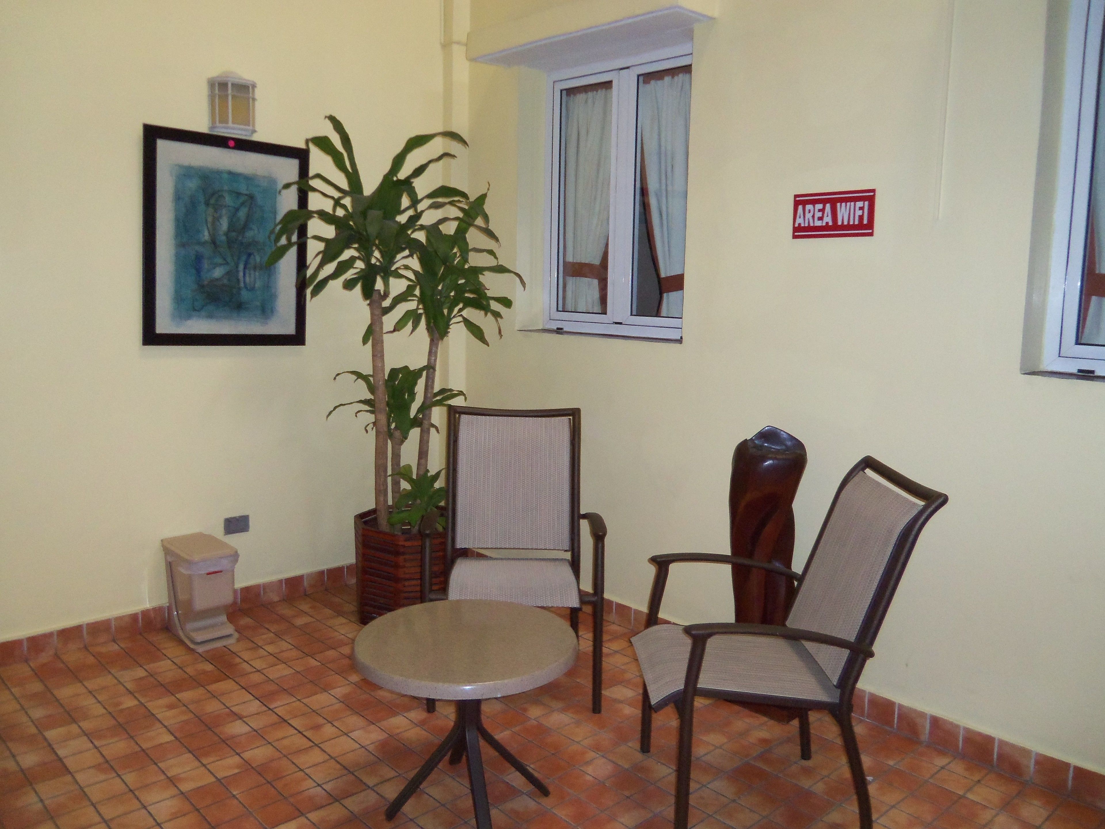 A small room with a tiled floor, a round table, two chairs, a potted plant, a framed picture on the wall, and a sign indicating an area with WiFi.