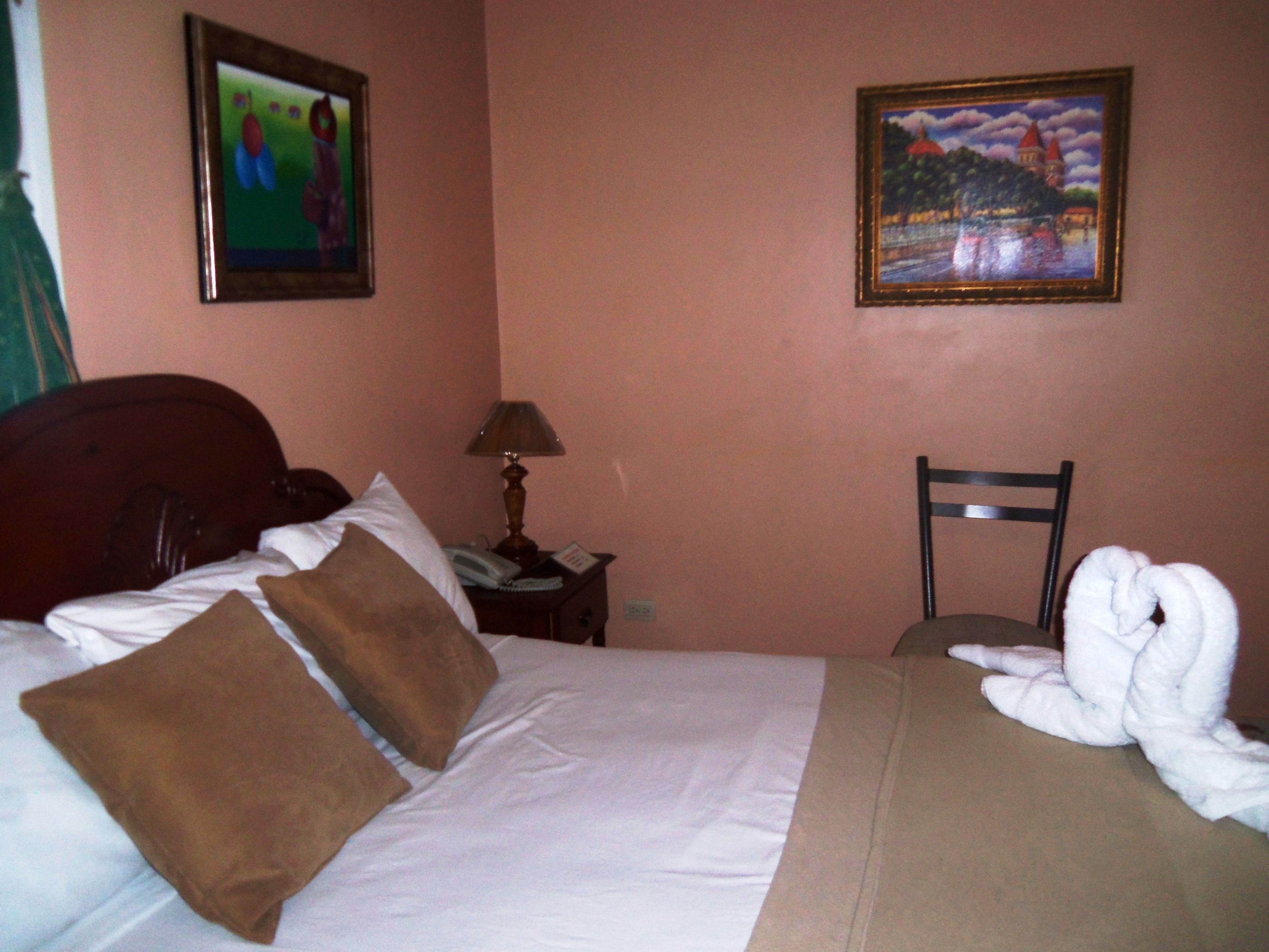 A hotel room with a bed, pillows, a chair, a nightstand with a lamp, and two framed pictures on the wall.