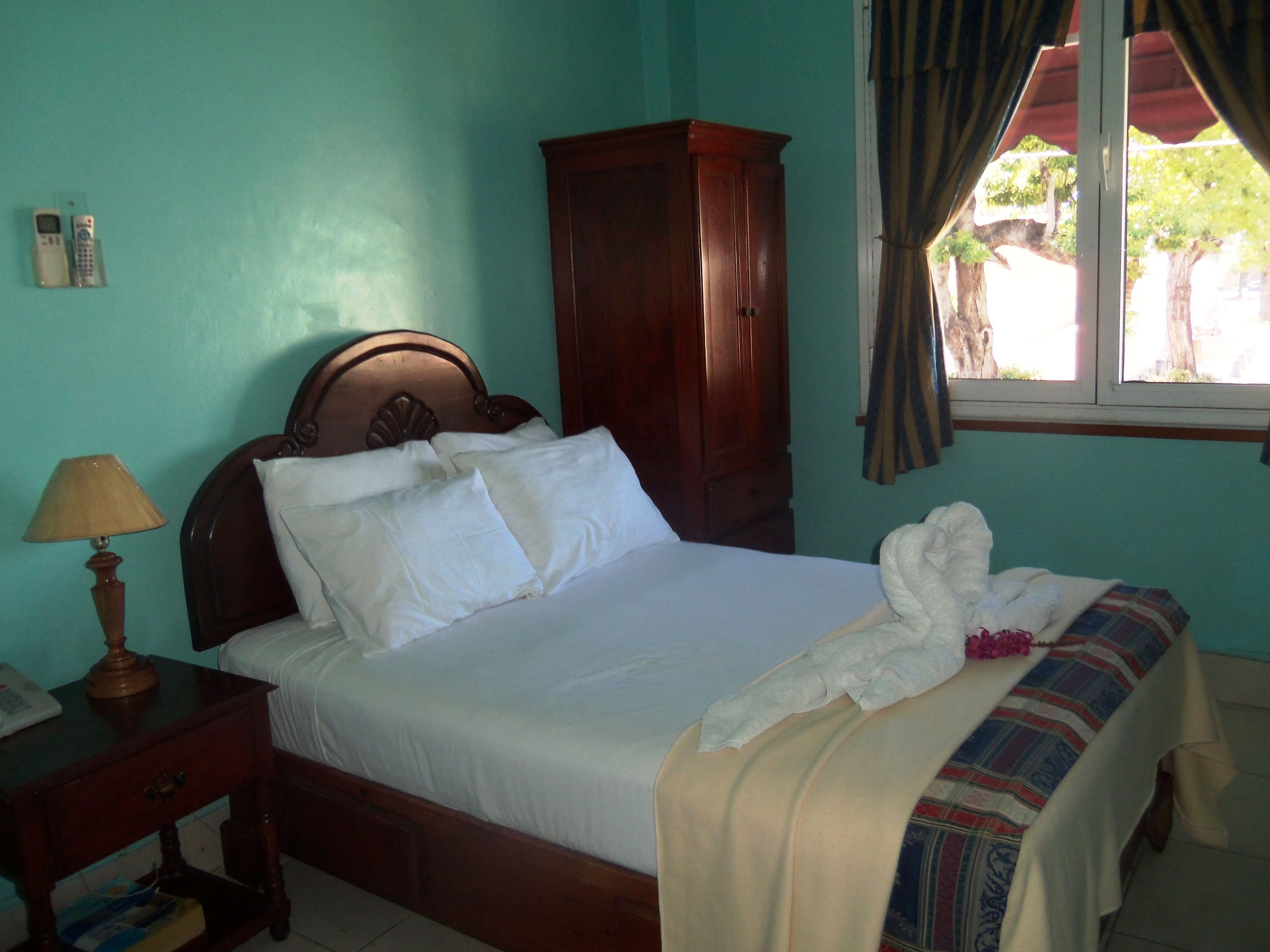 A neatly made bed with white linens, a wooden headboard, a wooden wardrobe, a bedside lamp, and a window with curtains.