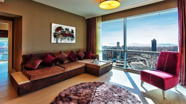 Un salon moderne avec un grand canapé, un fauteuil rouge et une vue sur la ville à travers de grandes fenêtres.