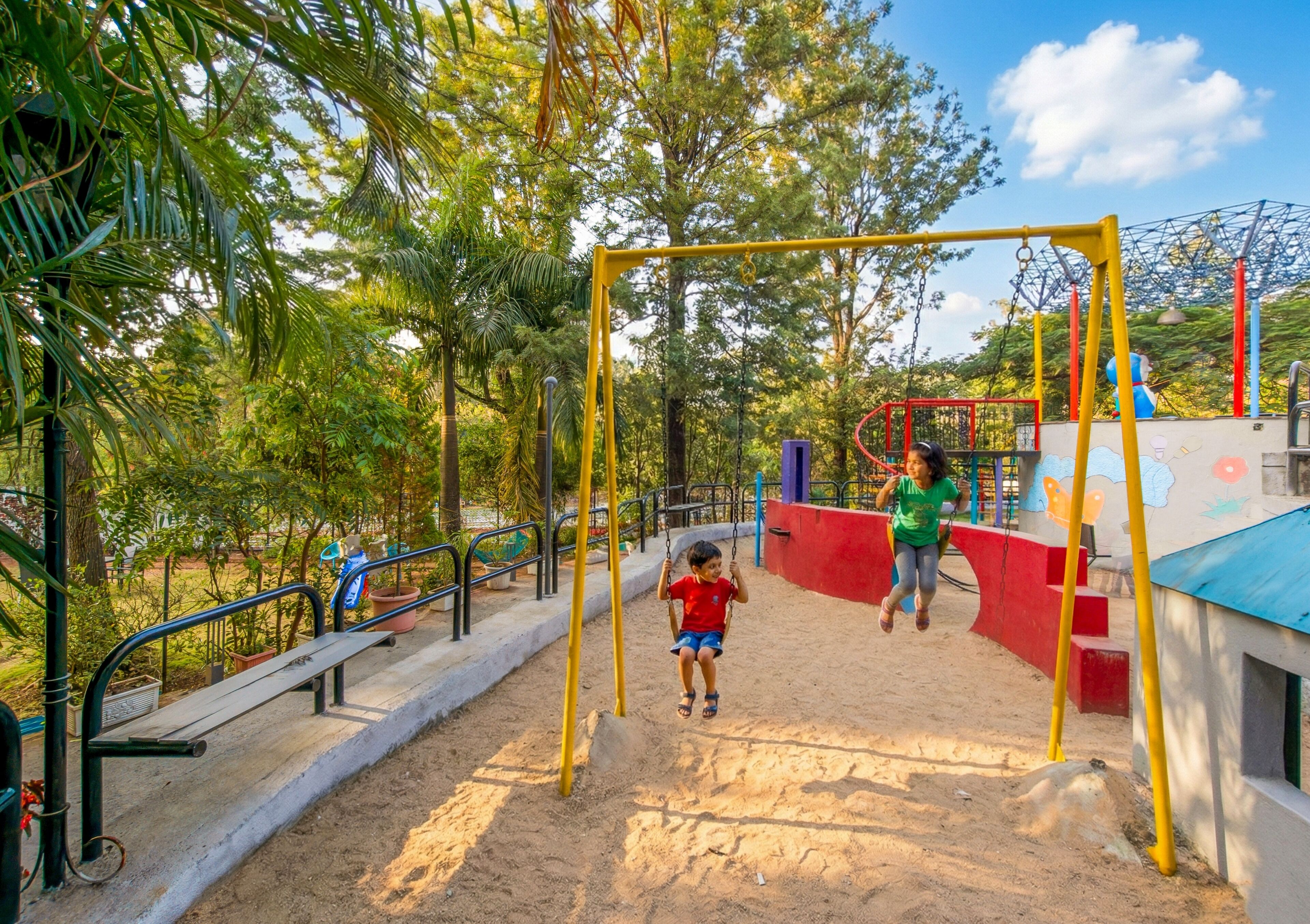 Ein Spielplatz mit Schaukeln und Sandkasten, umgeben von Bäumen und blauem Himmel.