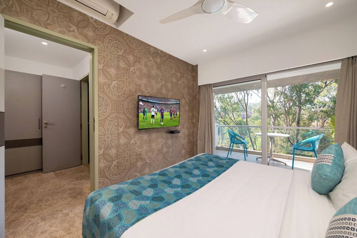 A hotel room with a bed, a TV, a balcony with chairs, and a view of trees.
