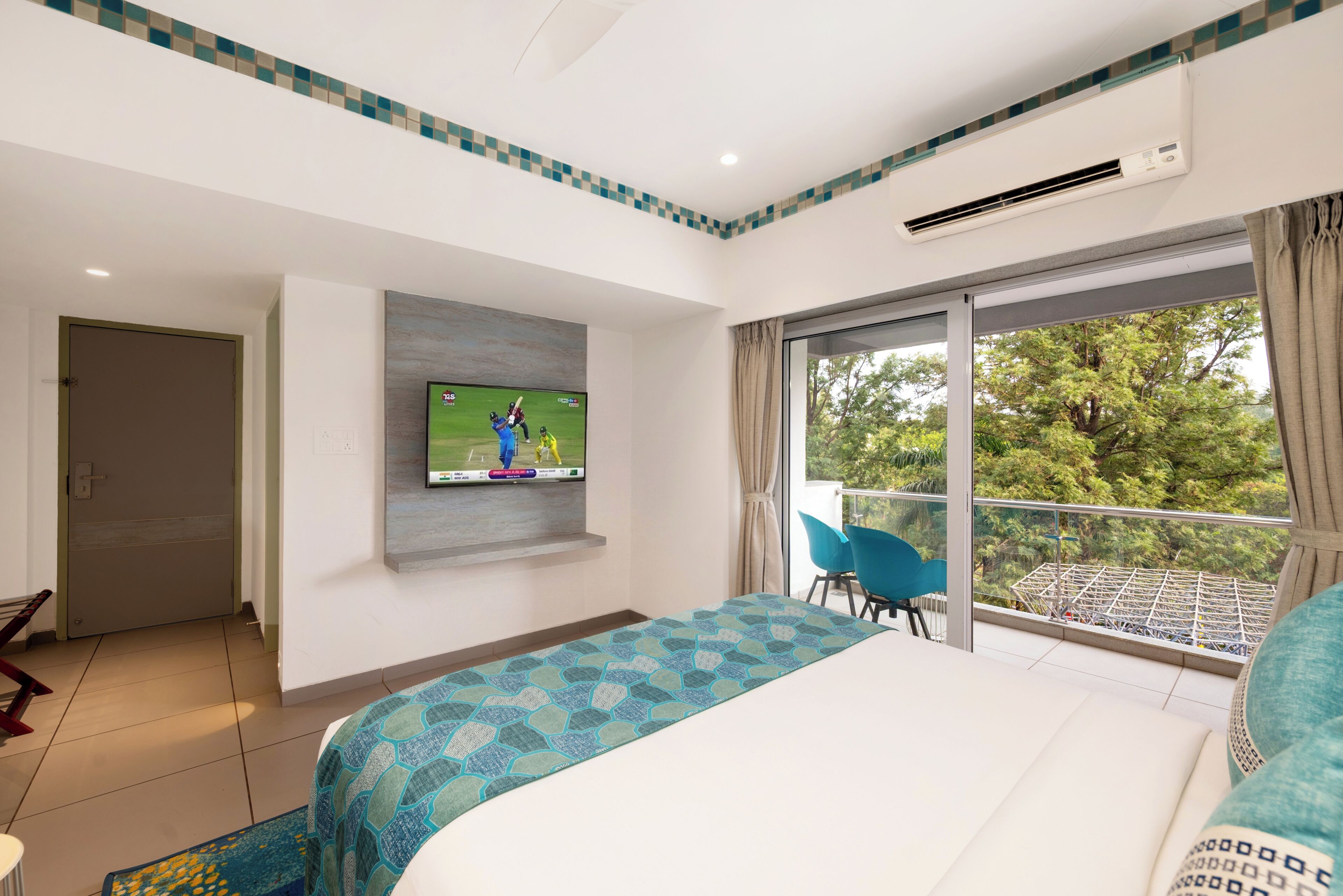 Hotelkamer met een groot bed, een flatscreen televisie aan de muur, een balkon met uitzicht op bomen en een raam met gordijnen.