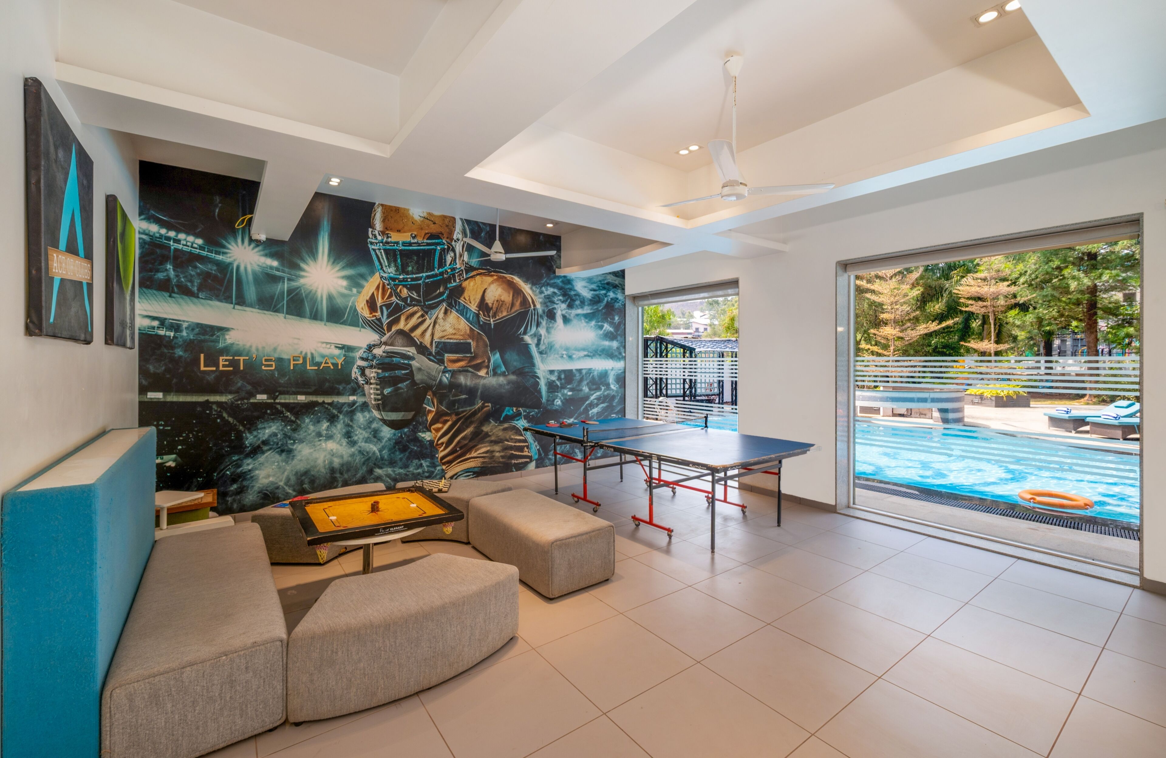 A modern indoor space with a ping pong table, comfortable seating, a mural, and a view of an outdoor swimming pool.