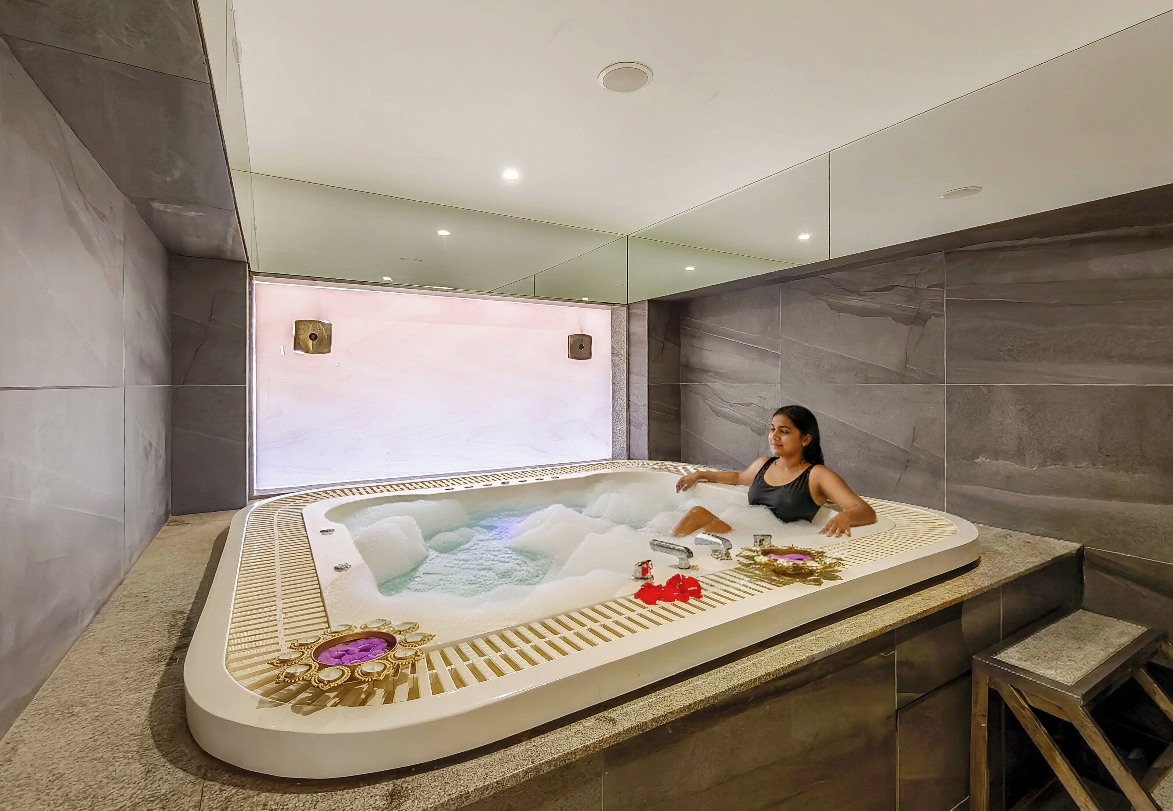 A person relaxing in a modern, freestanding hot tub with a built-in massage feature, surrounded by a minimalist bathroom with a large mirror and a view of the outdoors.