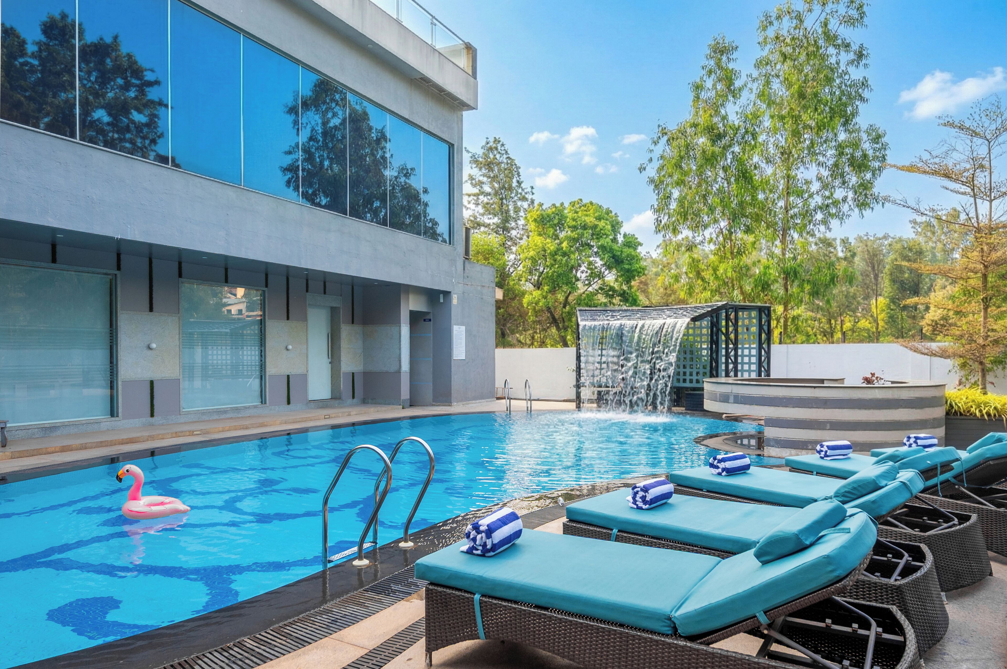 Una piscina con sillones de descanso, un elemento acuático y un edificio moderno con amplios ventanales.