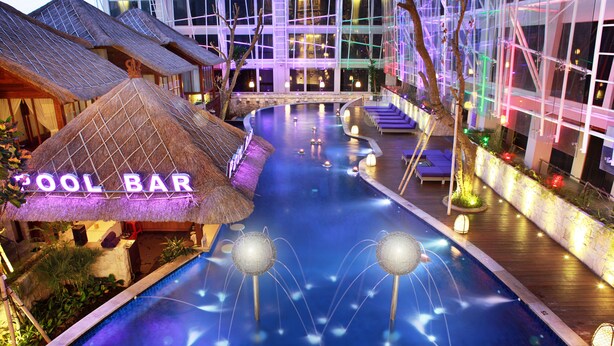 A swimming pool area with a bar named "Cool Bar," surrounded by thatched-roof huts and modern buildings with illuminated interiors. - Grand Mega Resort & Spa Bali (Kuta)