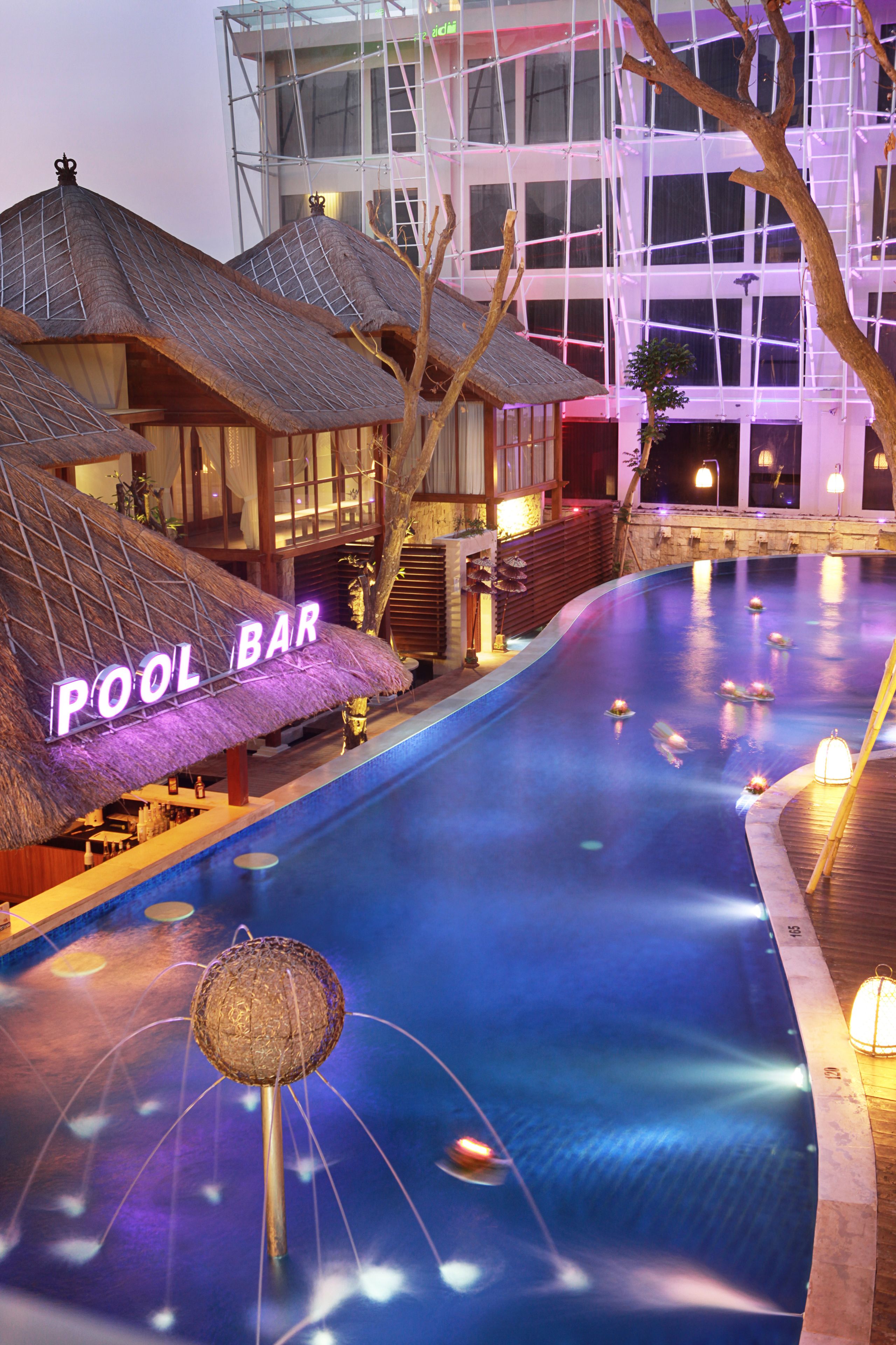 Zona de piscina con un letrero que dice 'POOL BAR', un área de bar con techo de paja y un edificio moderno al fondo.
